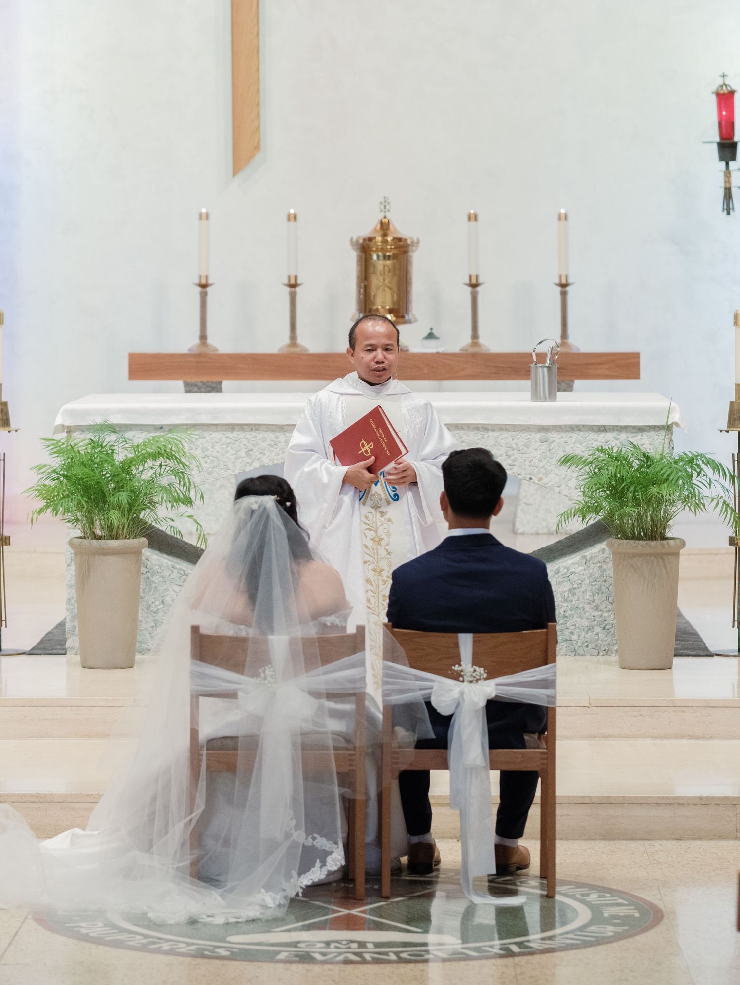 A wedding ceremony takes place in a modern church with potted plants and a wooden cross on the altar.