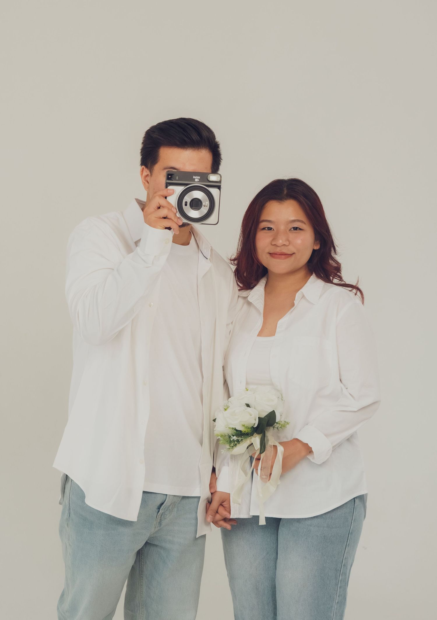Couple in white shirts and jeans takes creative photos against neutral background.