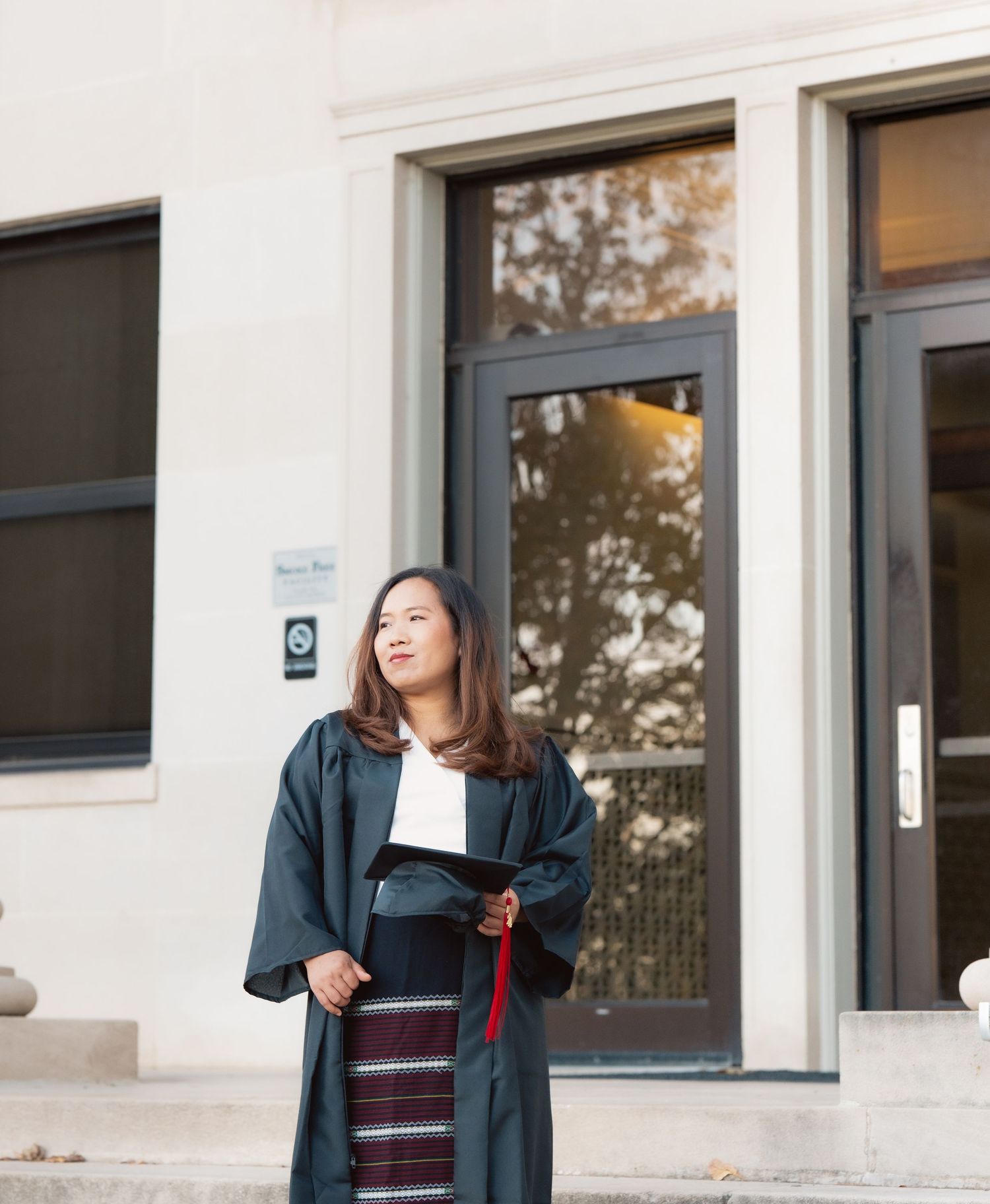 Graduate in academic regalia posing outside modern university building windows.