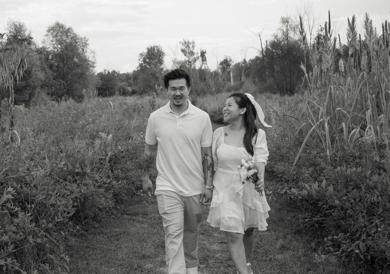 A black and white photo of a couple walking together on a dirt path through a meadow.