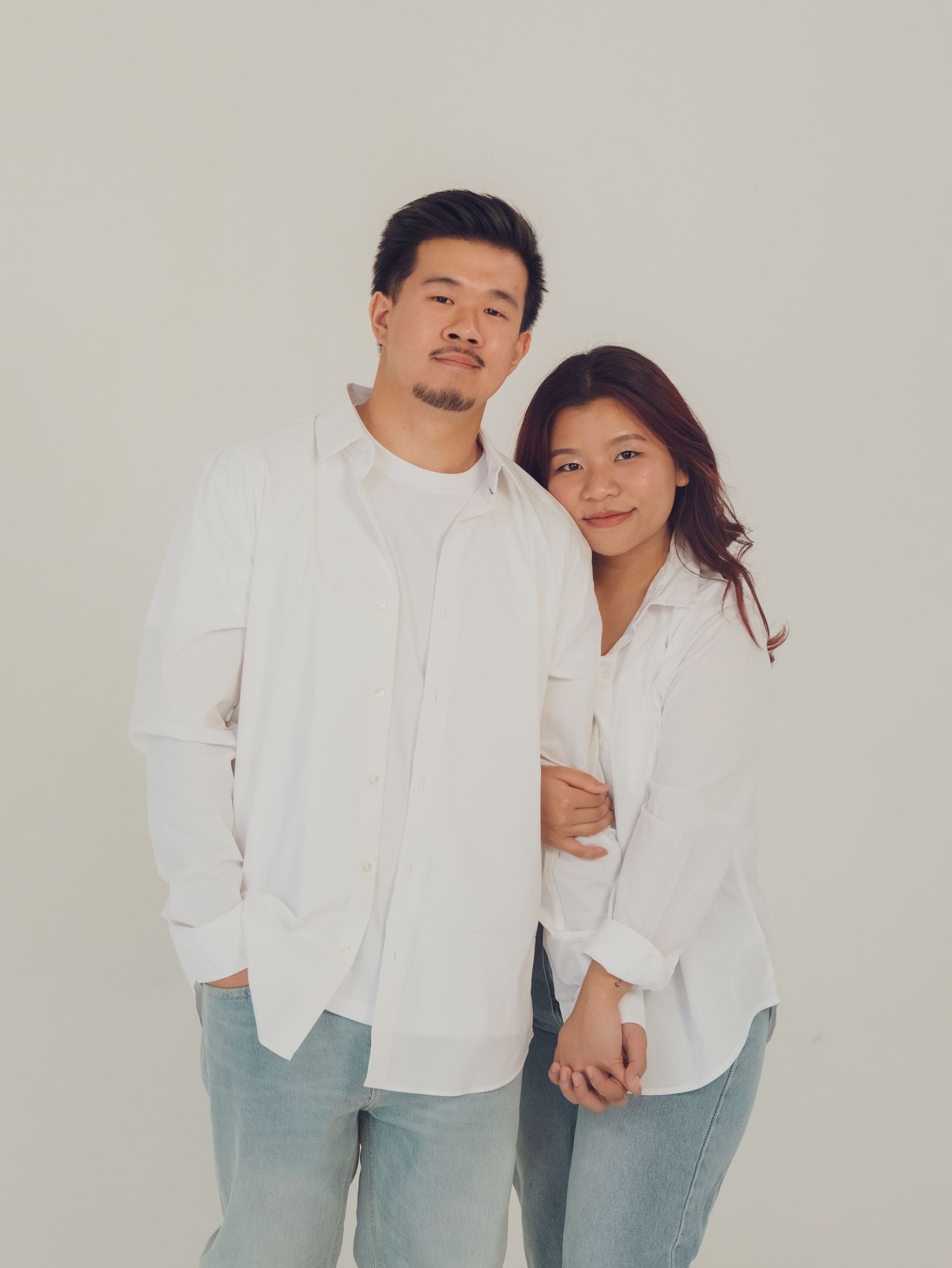 Couple in matching white button-down shirts and light denim jeans pose playfully against neutral background.
