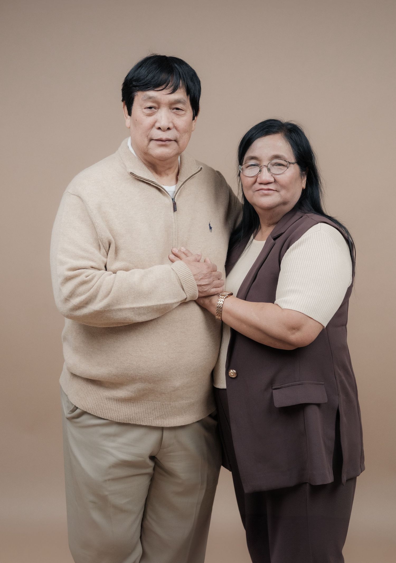 A couple standing close together wearing matching neutral colors against a brown backdrop.