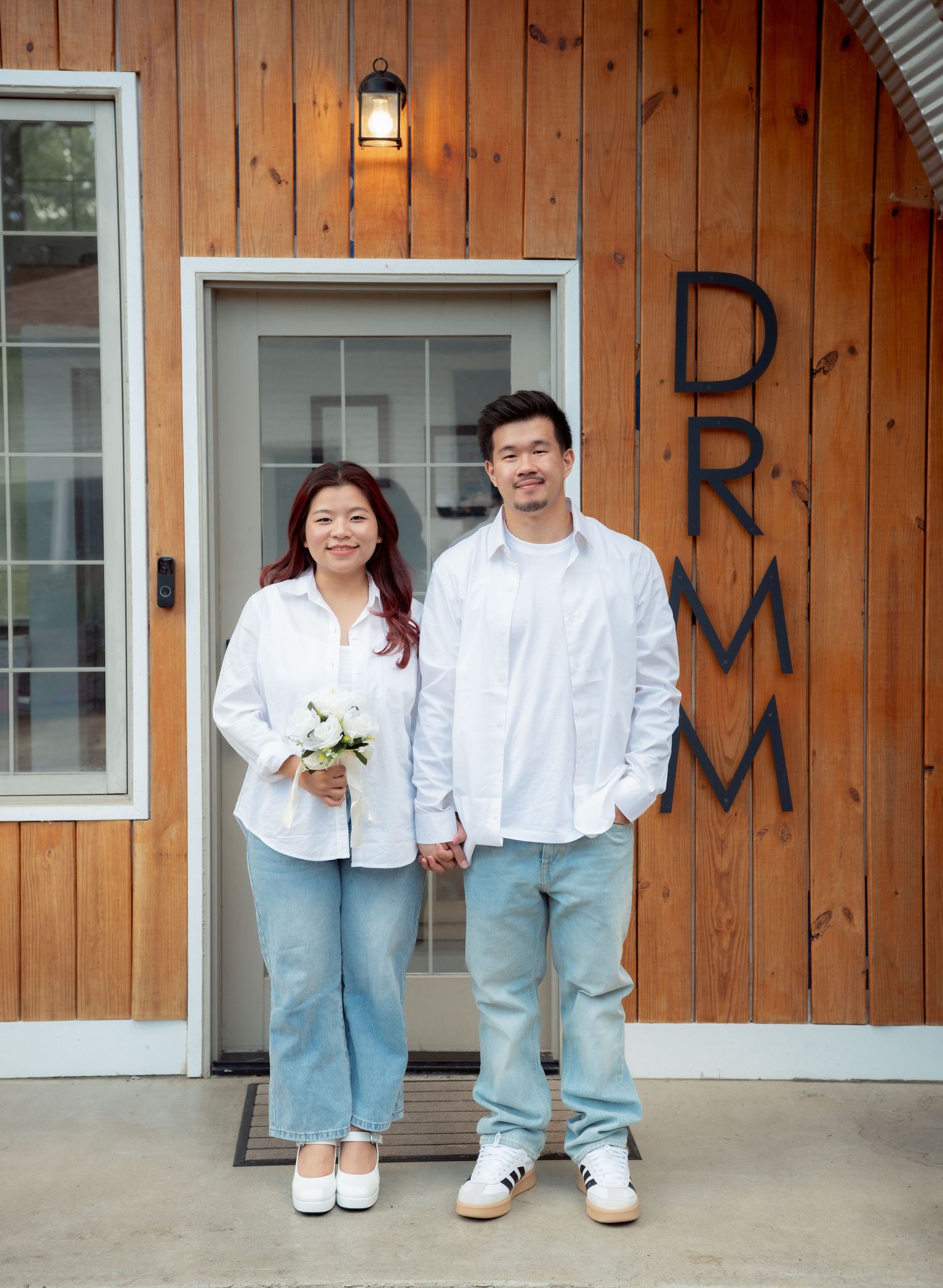 A couple in matching white tops and light denim poses against a wooden wall exterior.