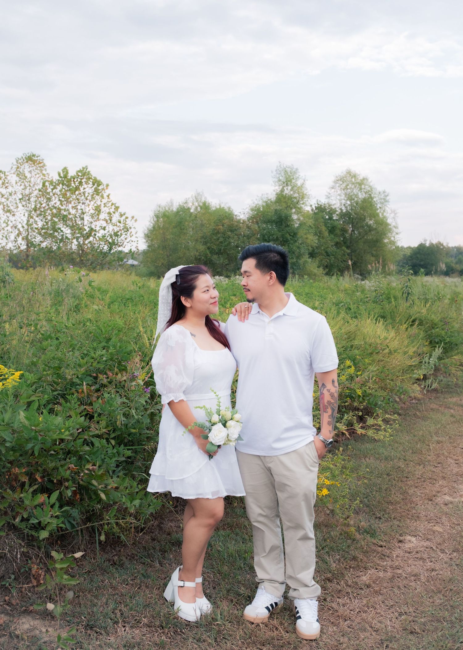 A couple in white outfits shares a romantic moment on a nature path with flowers.