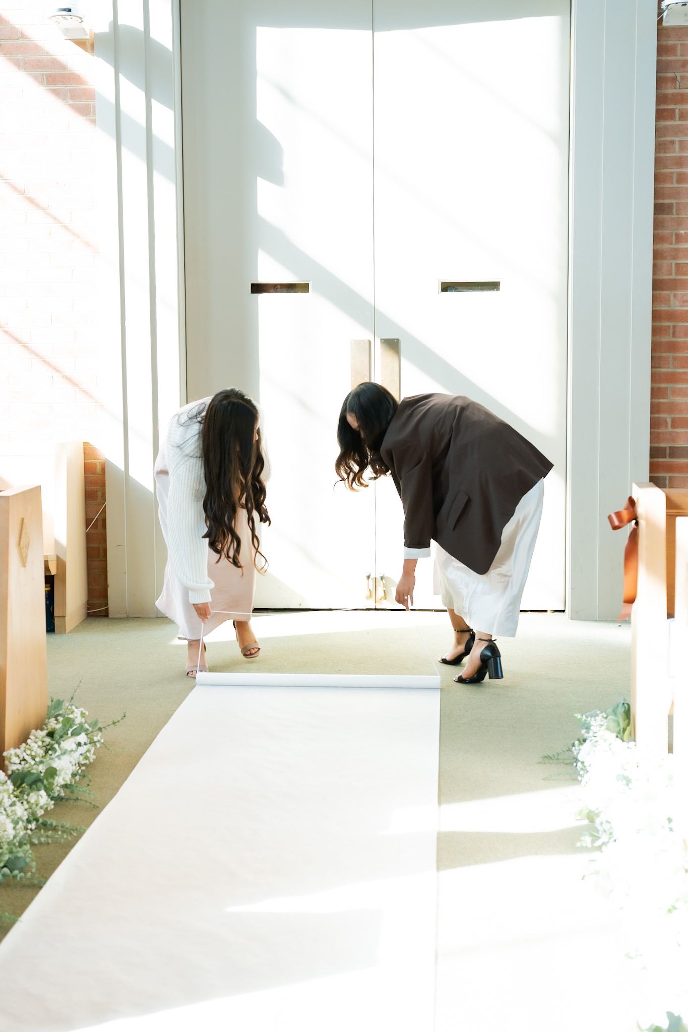 Two women unrolling a white aisle runner in a sunlit church before a wedding ceremony.