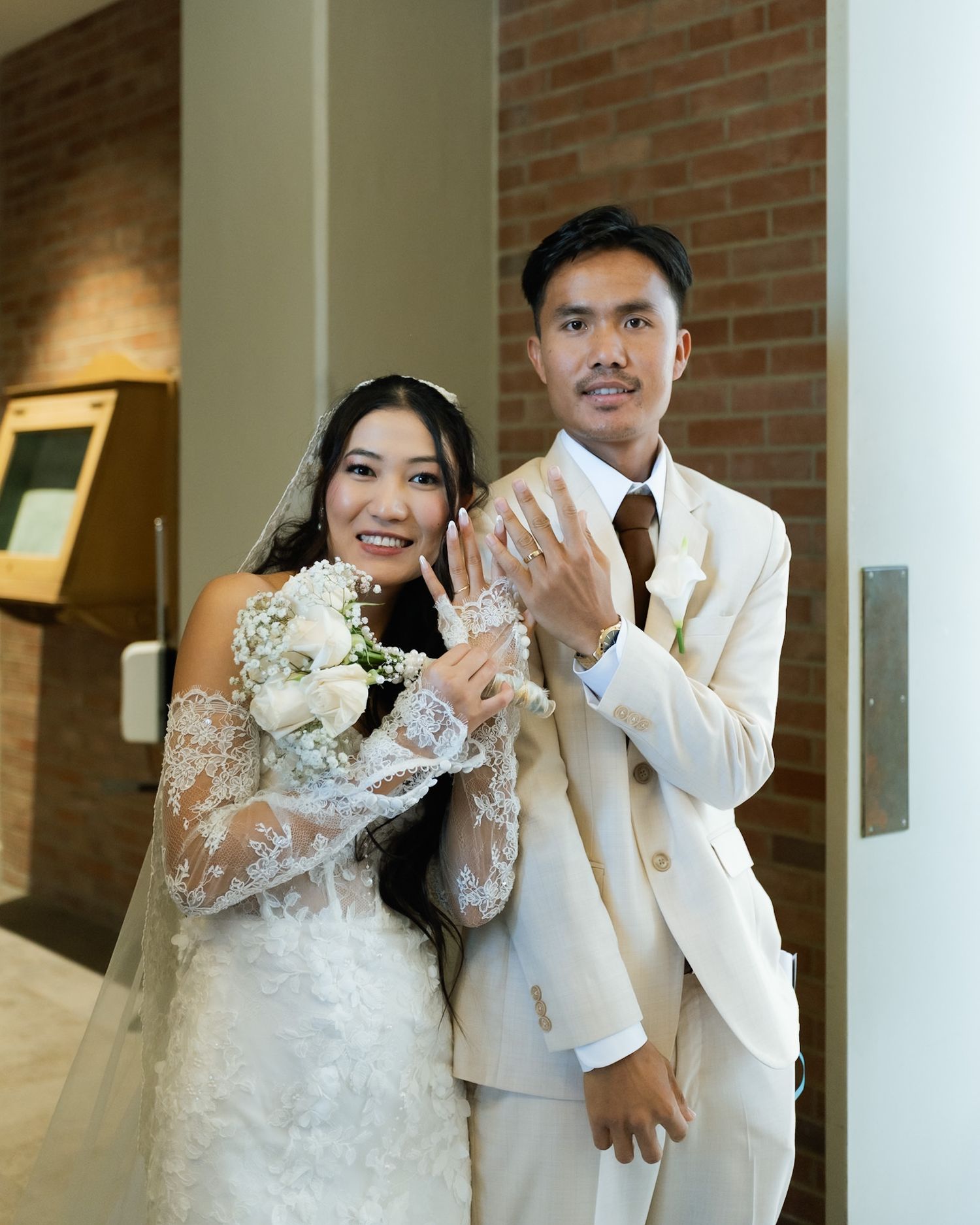 Newlywed Asian couple in wedding attire smiling and showing off wedding rings, bride holding white bouquet.
