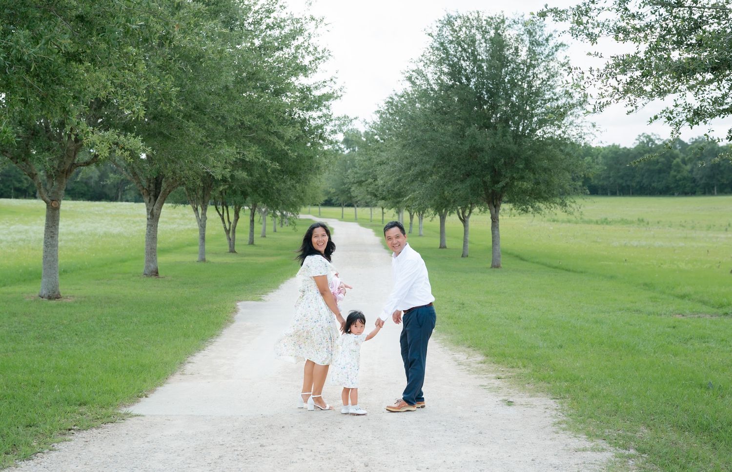 Family of three posing on a tree-lined gravel path in a lush green park, parents holding toddler's hands.