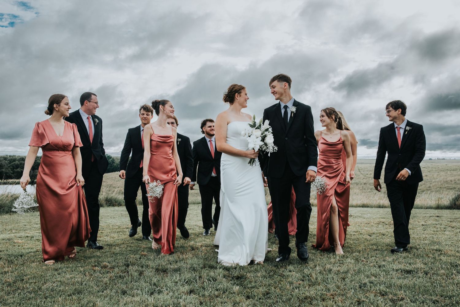 Wedding party walks outdoors on green grass, bride in white gown with bridesmaids in pink dresses and groomsmen in black suits.