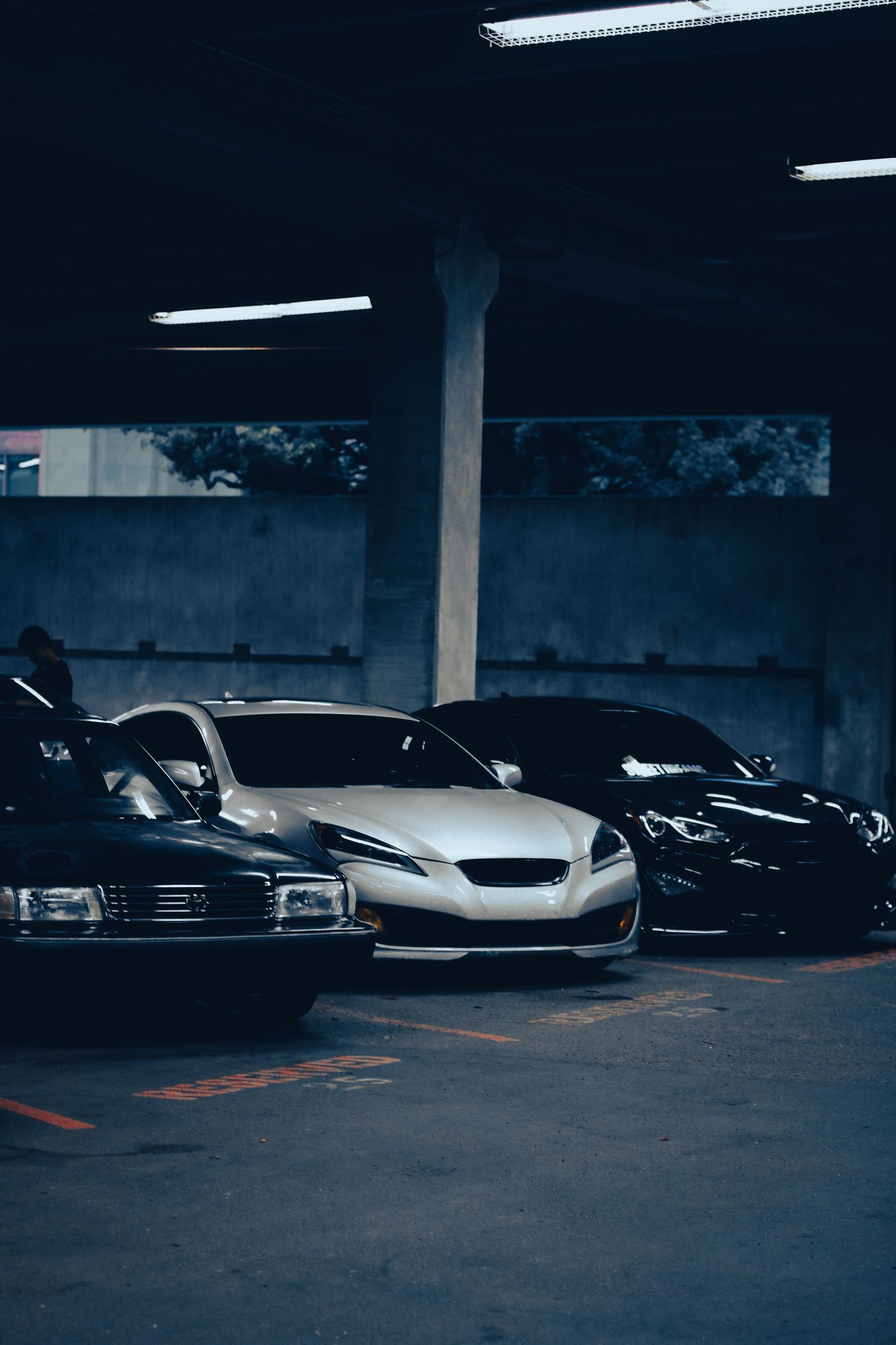 Three sleek cars parked in a dimly lit parking garage, featuring a white sports car flanked by two dark vehicles.