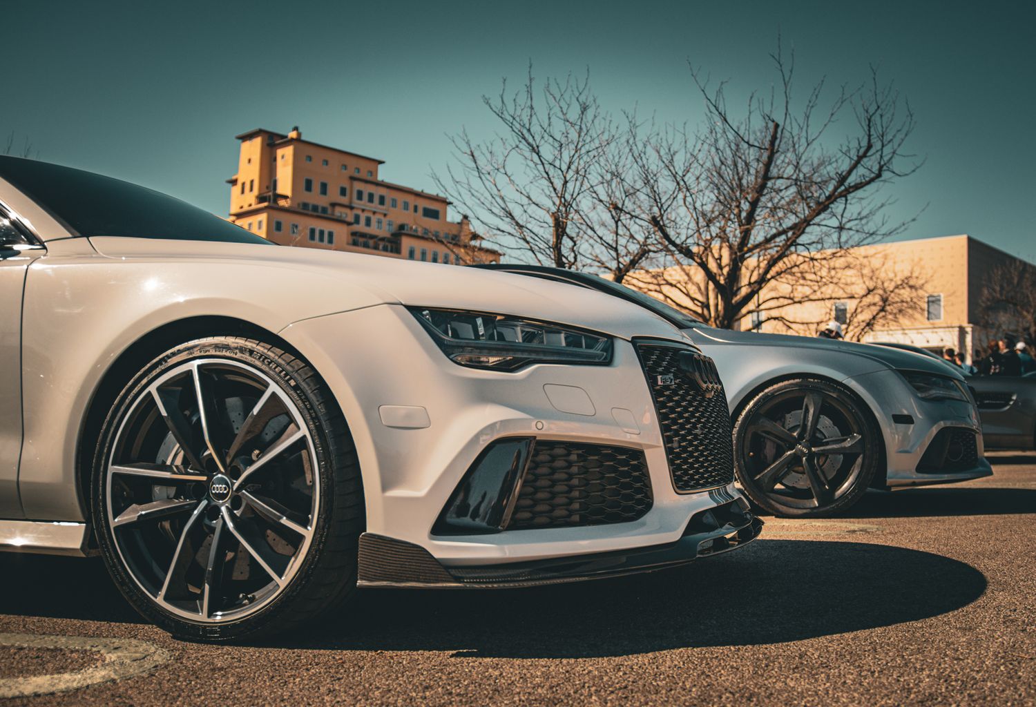 Two white Audi RS7 sports cars parked side by side in a sunny outdoor lot with bare trees.