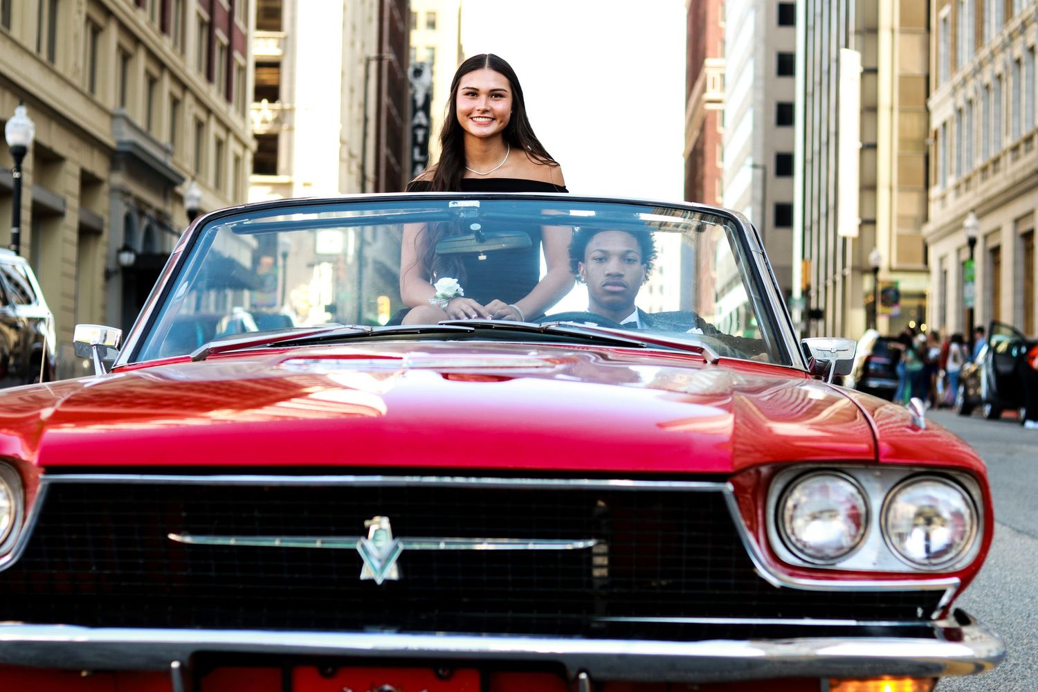 Two people riding in a classic red convertible car on a city street on a sunny day.