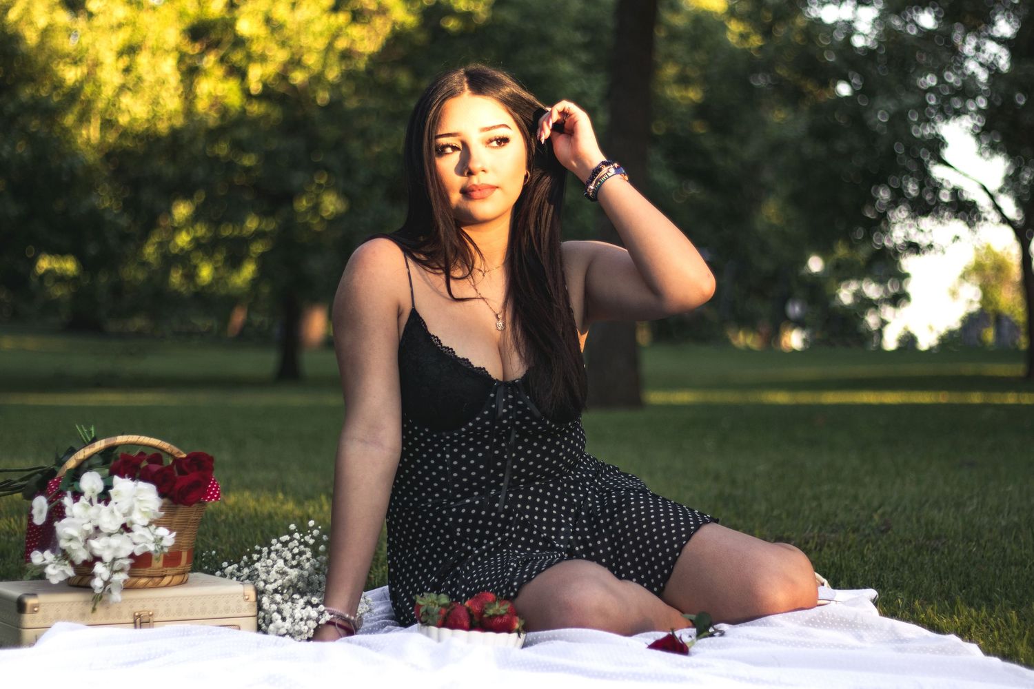 Young woman in black polka dot dress sitting on white blanket in park with flower basket and roses.