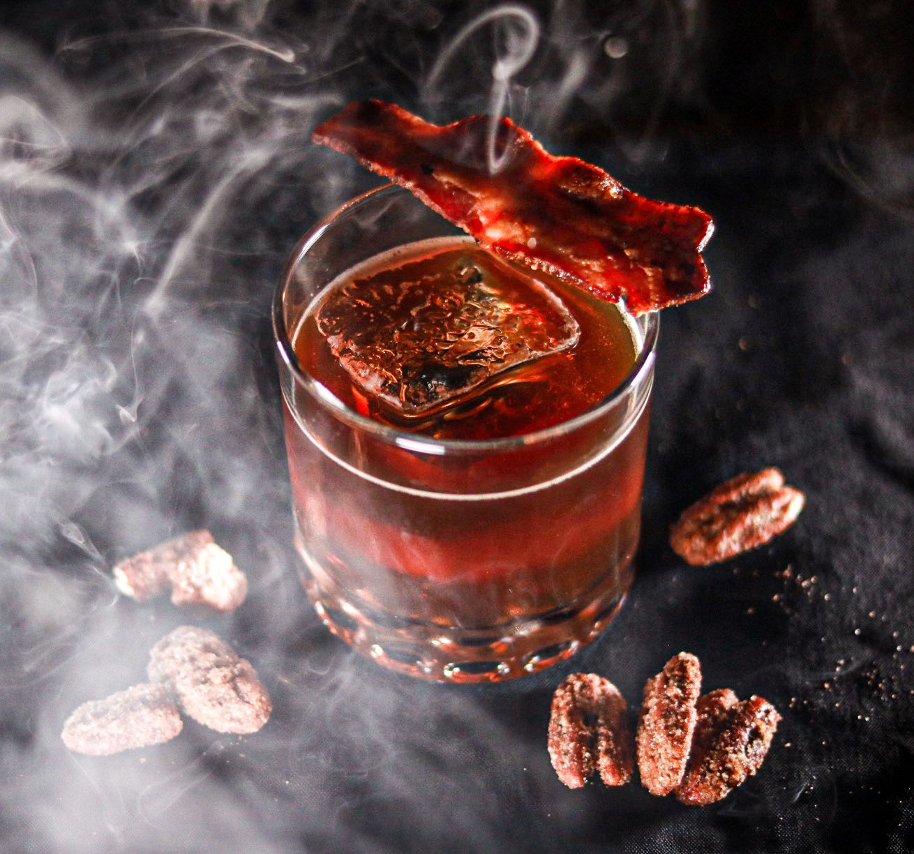 Smoky bourbon cocktail in a rocks glass garnished with crispy bacon strips, surrounded by candied pecans on a dark background.