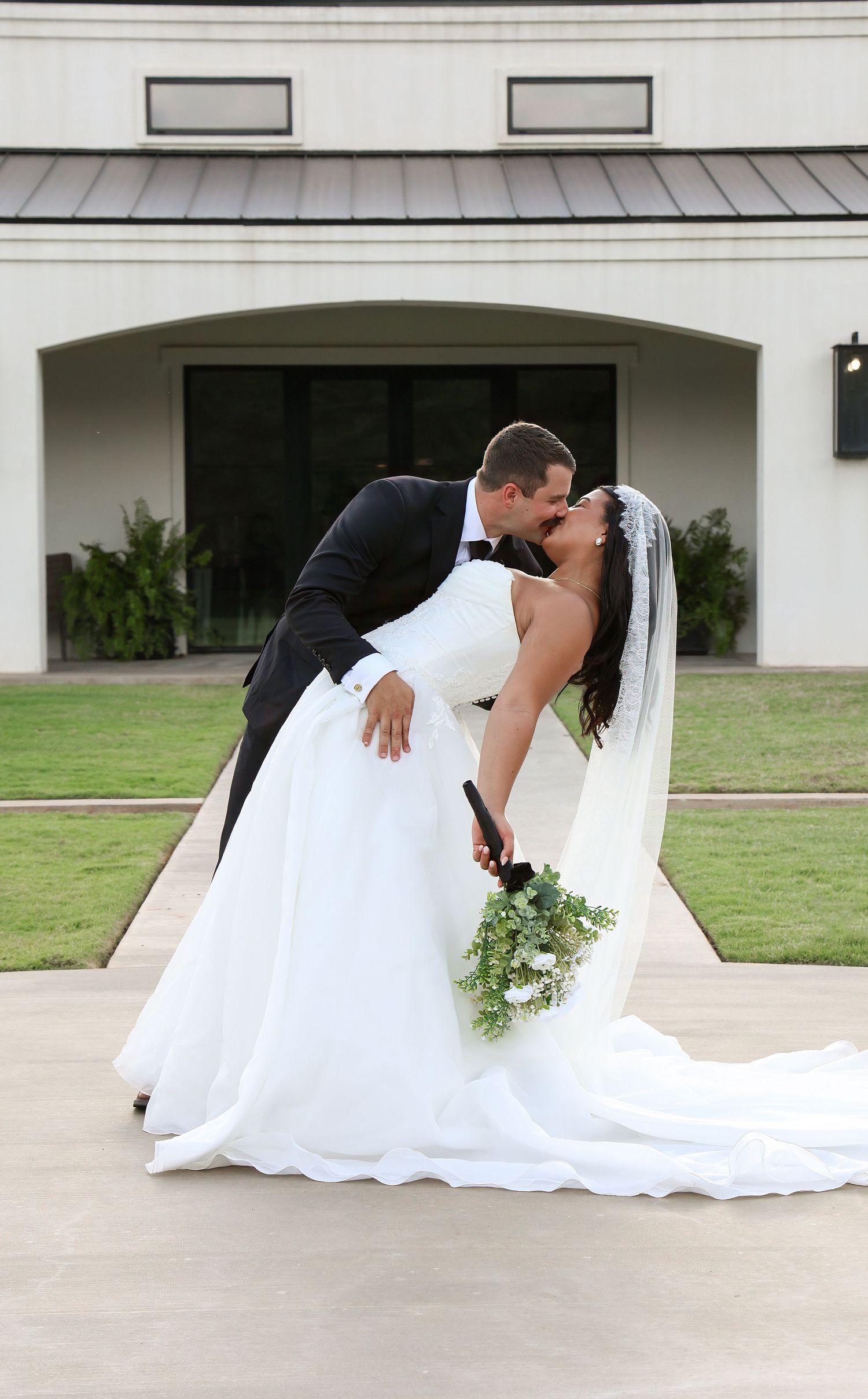 Bride and groom share a romantic dip kiss in front of an elegant white venue, bride holding a green and white bouquet.
