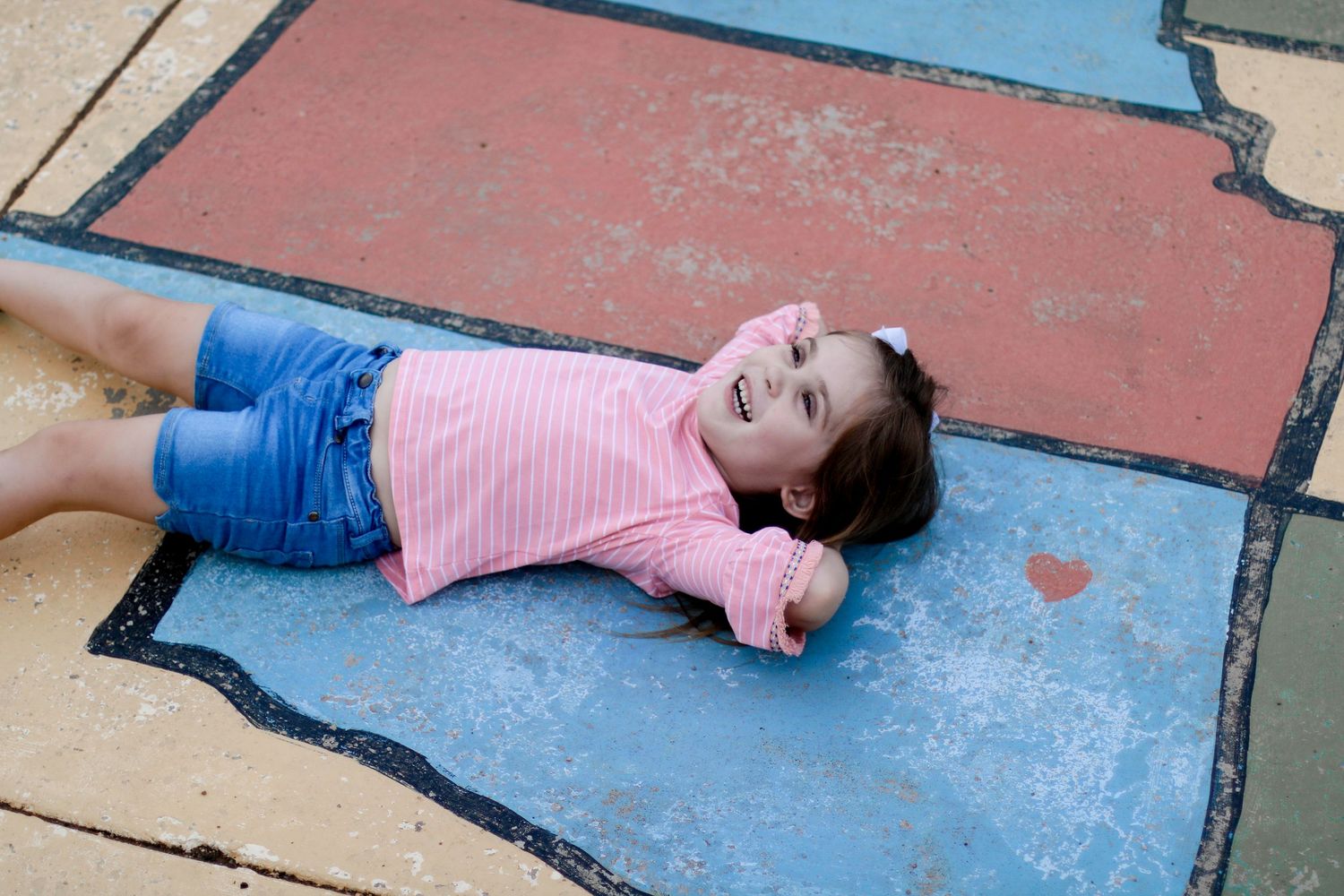 Someone wearing a pink striped shirt and blue shorts lies laughing on a painted concrete surface outdoors.