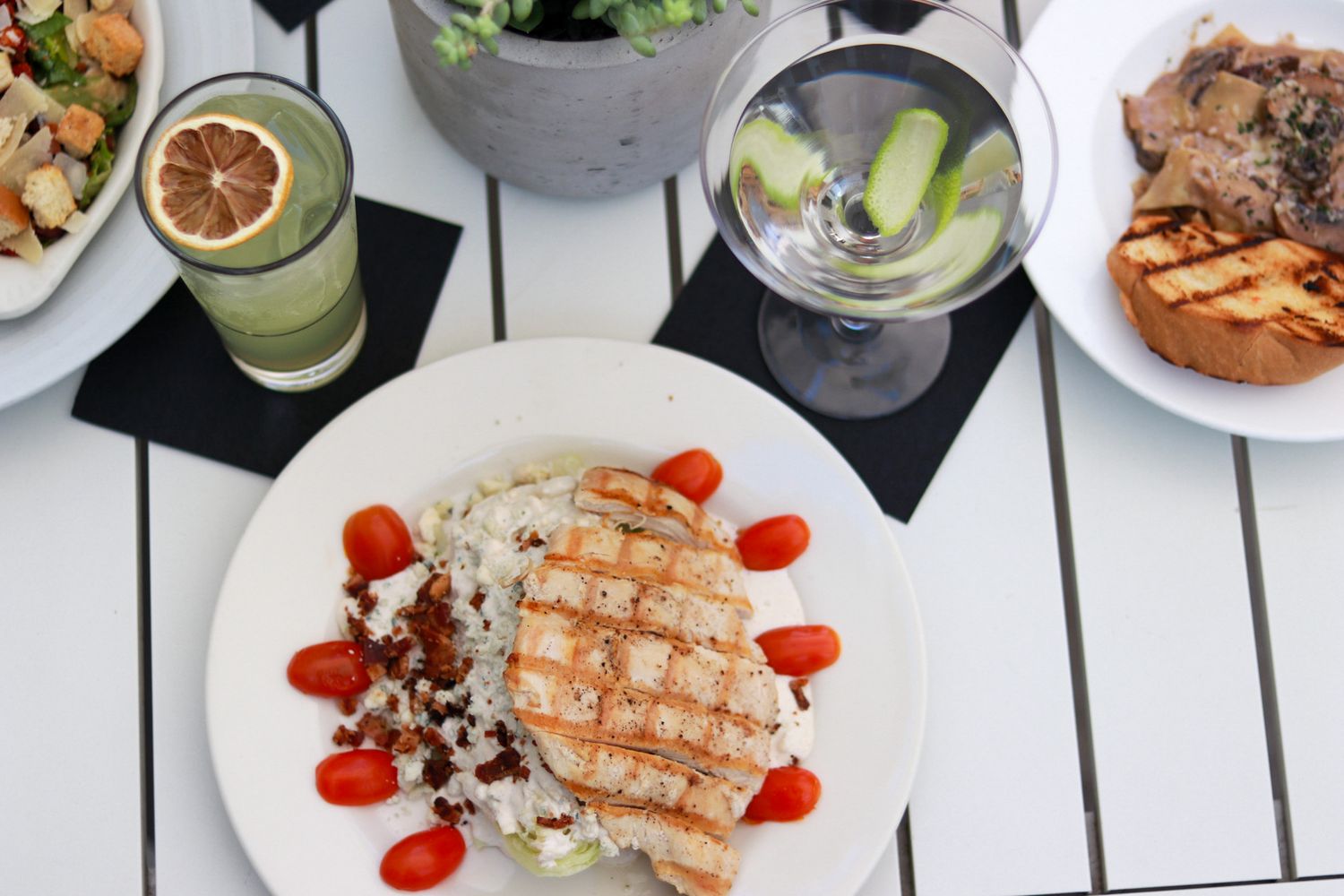 Grilled chicken breast with cherry tomatoes and rice on white plate, martini and green juice on outdoor dining table.