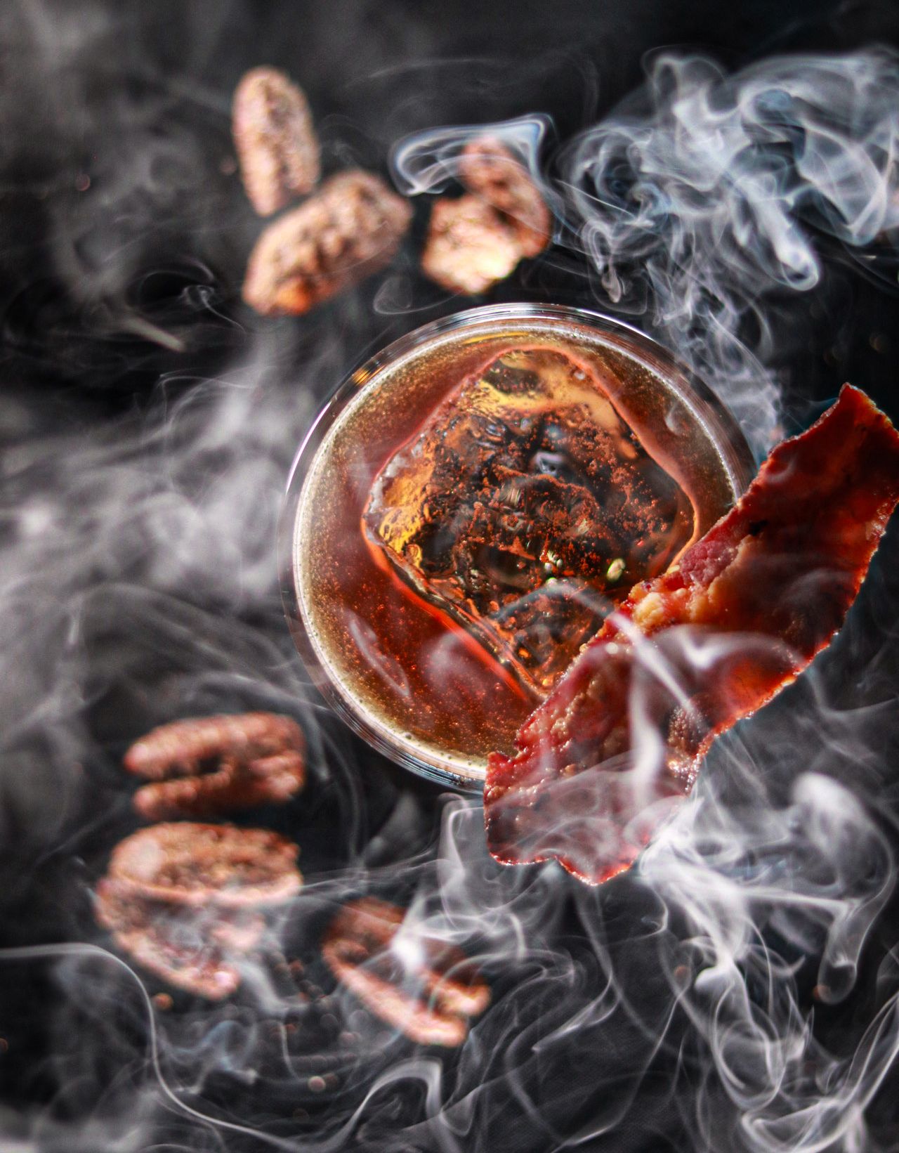 Smoky cocktail with bacon garnish, charred ice cube, and pecans floating in dramatic dark atmospheric photography.