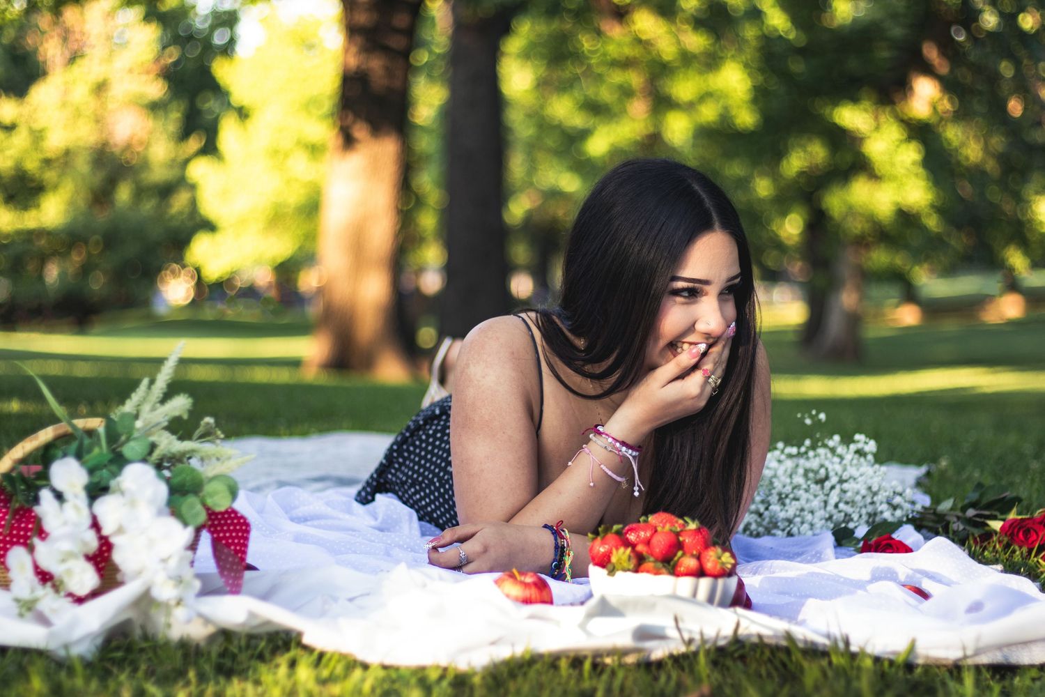 Young woman laughing on a white picnic blanket in a park, surrounded by strawberries and flowers on a sunny day.