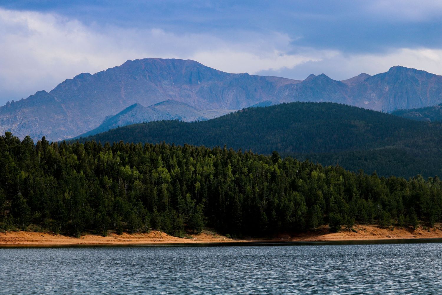 A serene mountain lake with dense evergreen forests and dramatic rocky peaks under a cloudy sky.