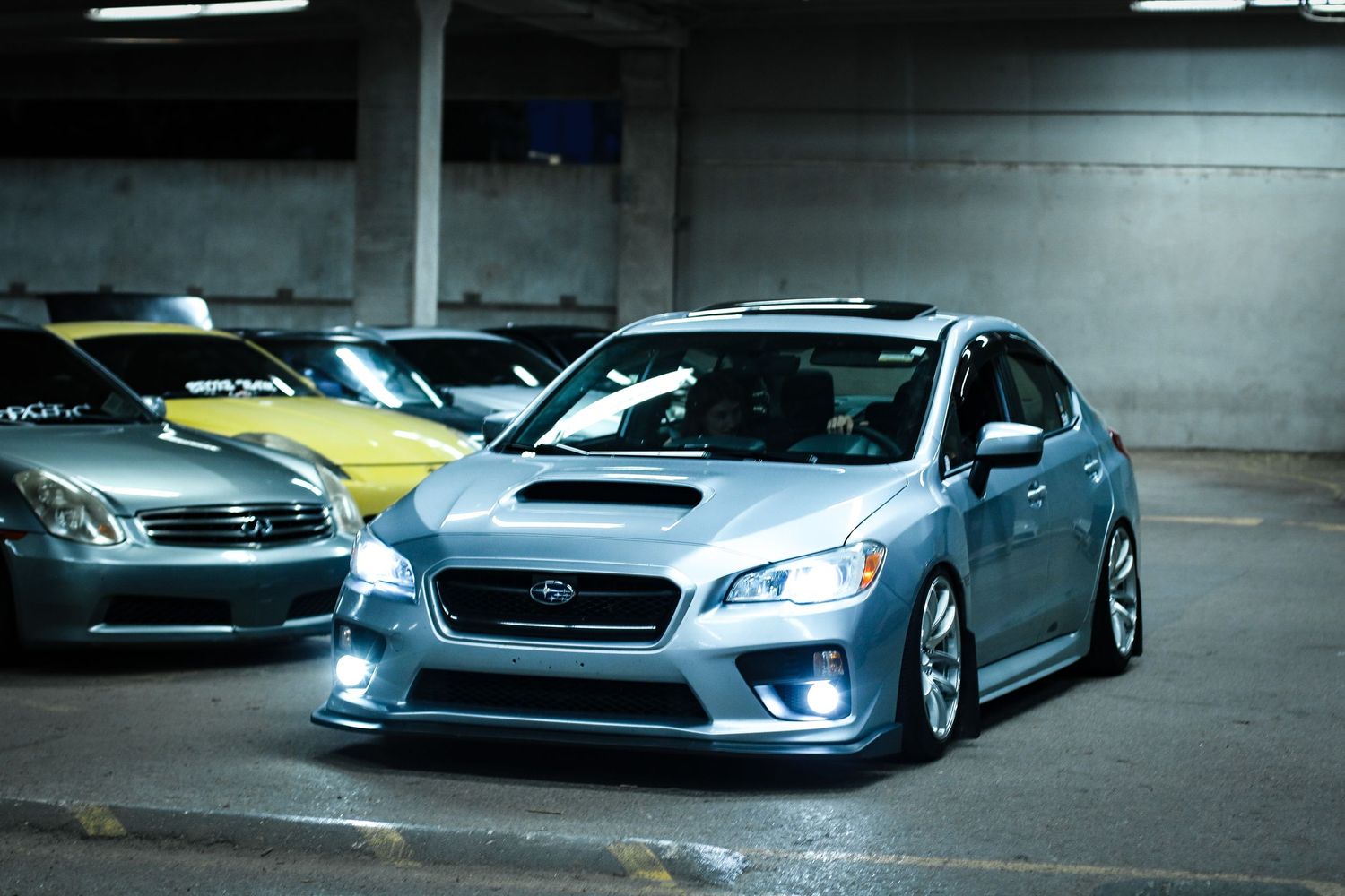 A lowered silver Subaru WRX parked in a garage with yellow and gray cars visible in the background.