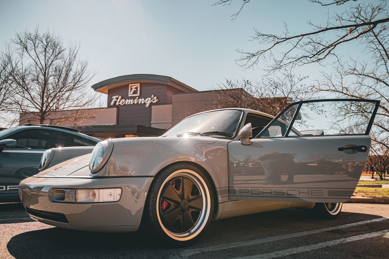Silver Porsche 911 parked outside Fleming's restaurant, featuring gold wheels and open door on a sunny day.