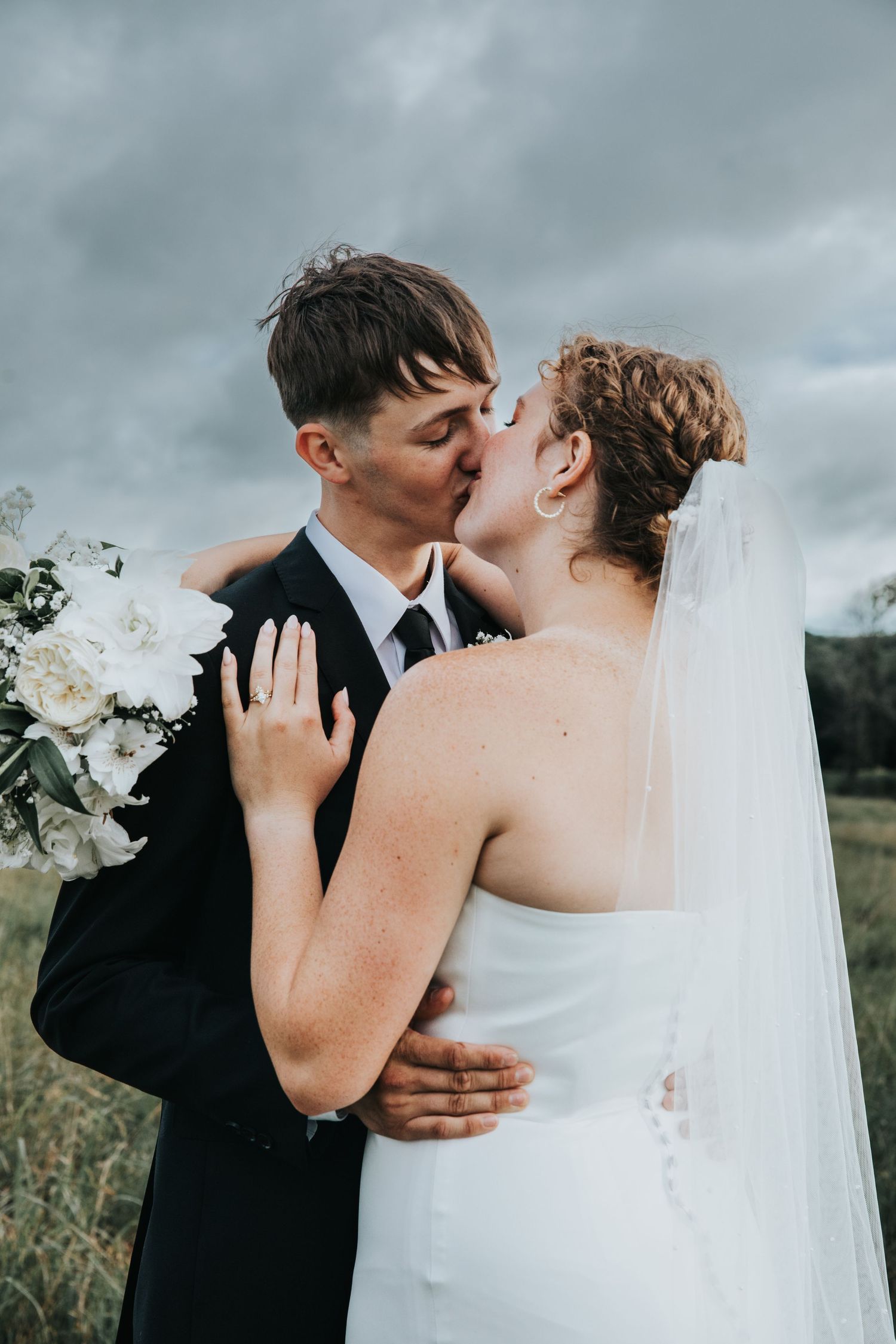 A bride and groom share a romantic kiss outdoors, with the bride holding white flowers and wearing a veil on a cloudy day.