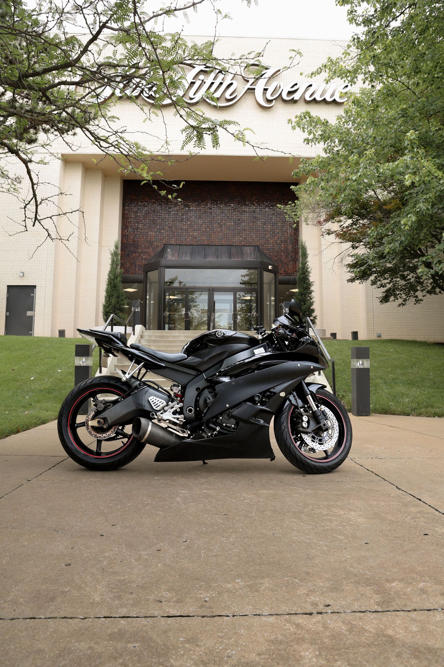 Black sport motorcycle parked in front of a Fifth Avenue building entrance with lush green trees and manicured landscaping.