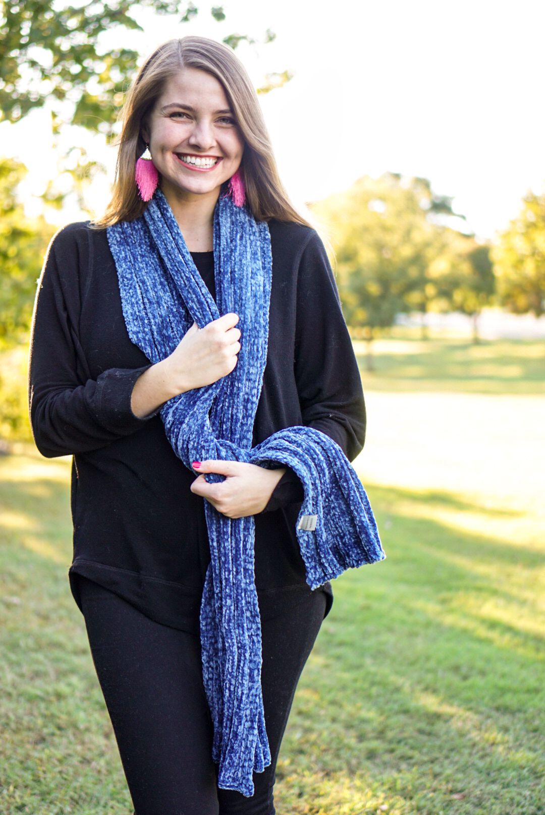 A person wearing a black sweater and blue scarf poses outdoors in a grassy field during golden hour lighting.