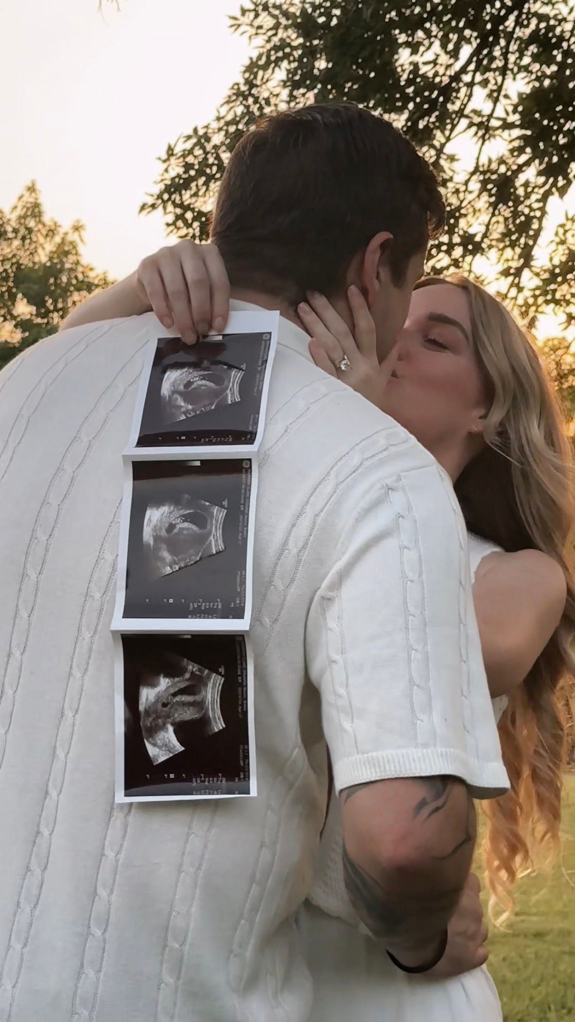 A couple embraces while holding ultrasound photos during sunset in a grassy field.