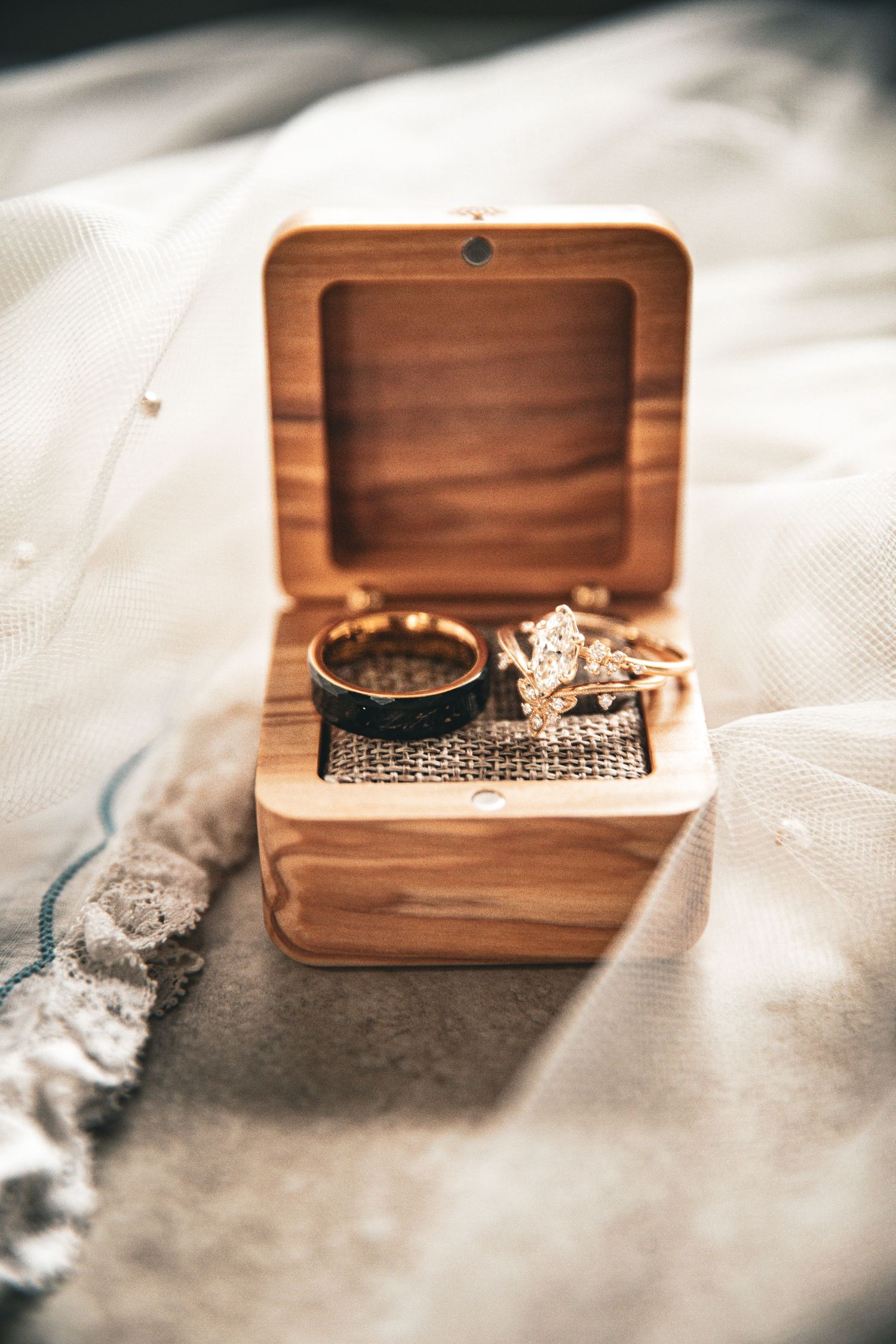 Gold wedding bands and diamond engagement ring displayed in an open wooden ring box on white bridal veil fabric.