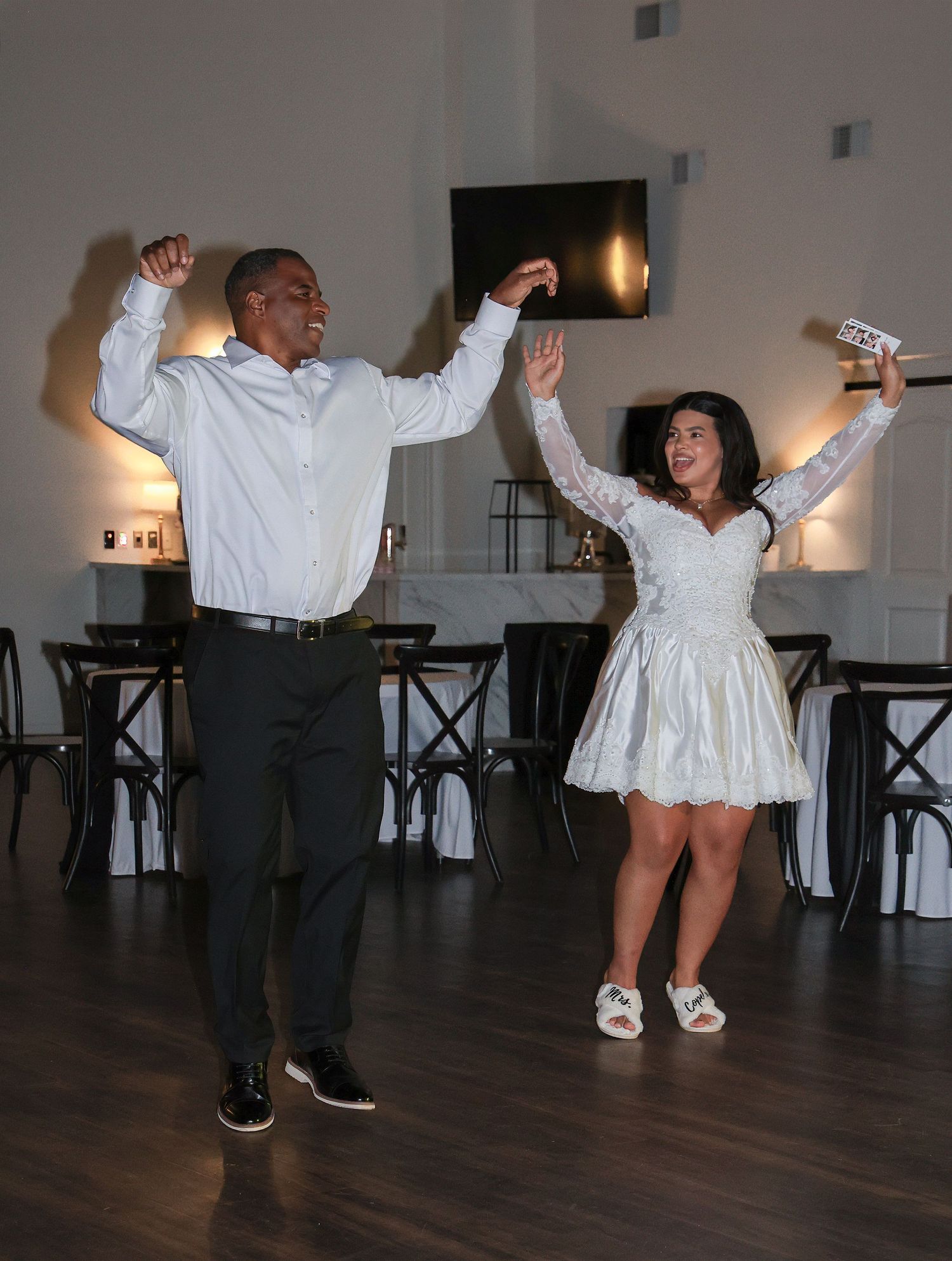 Couple dancing joyfully at wedding reception in white outfits in elegant venue.
