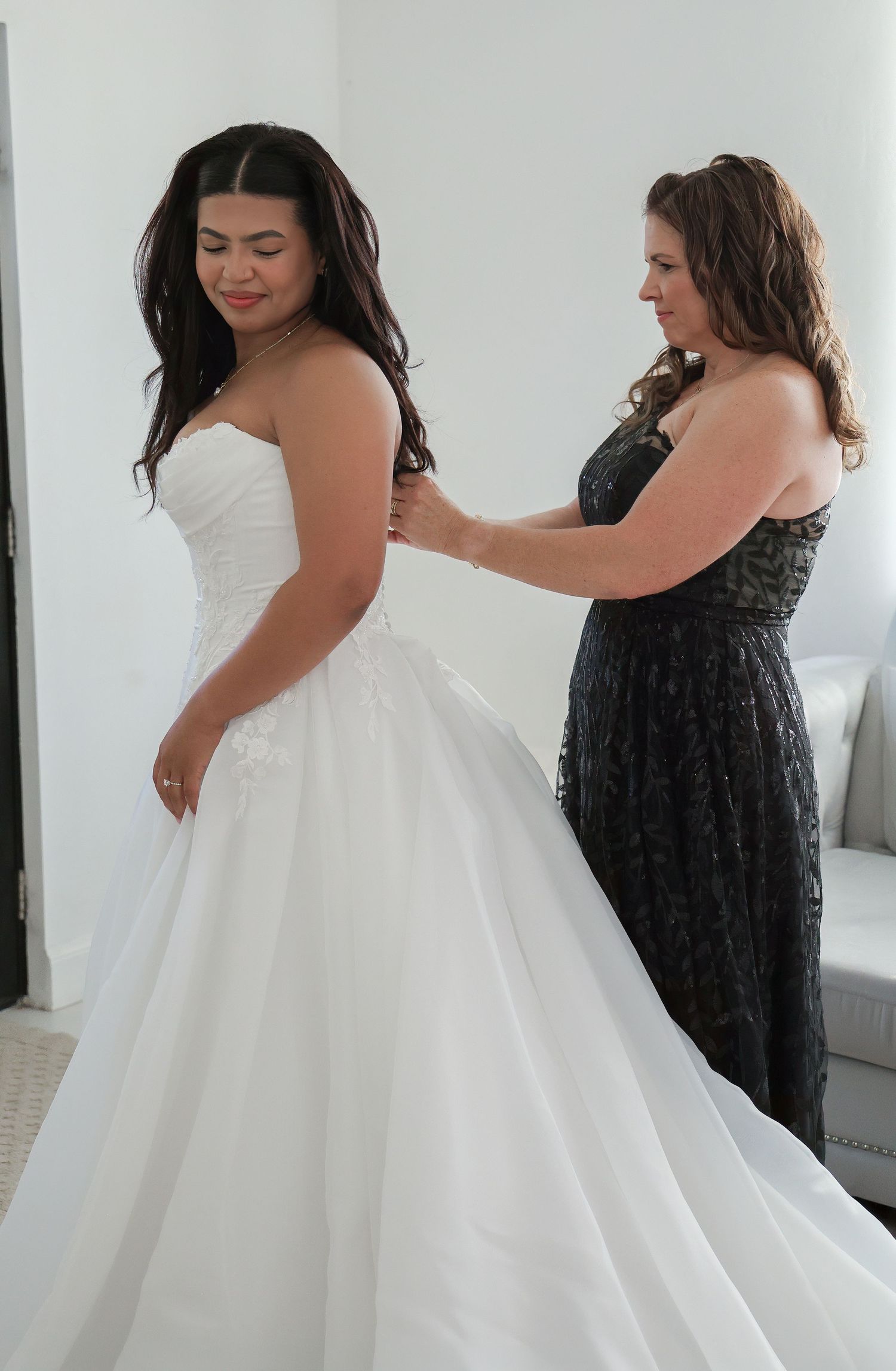 A woman in a white strapless wedding gown being helped by another woman in a black dress.