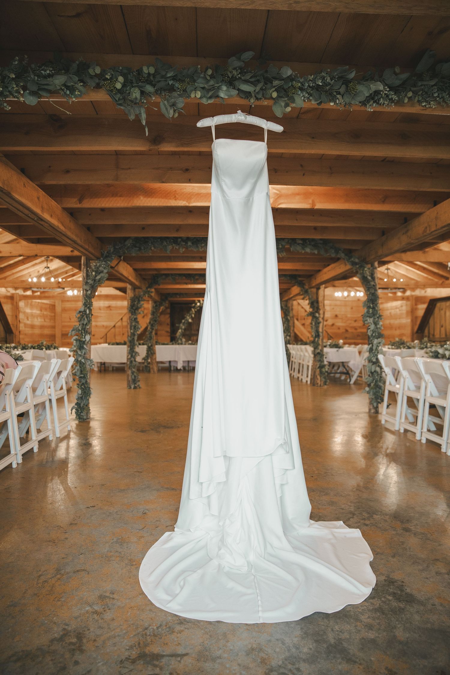 Elegant white wedding gown with long train hanging in a rustic barn venue decorated with greenery and white chairs.