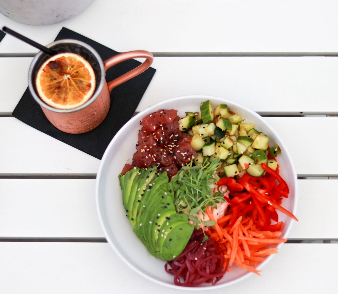 Colorful poke bowl with avocado, tuna, carrots, cucumber, and red pepper beside a copper mug with orange garnish.