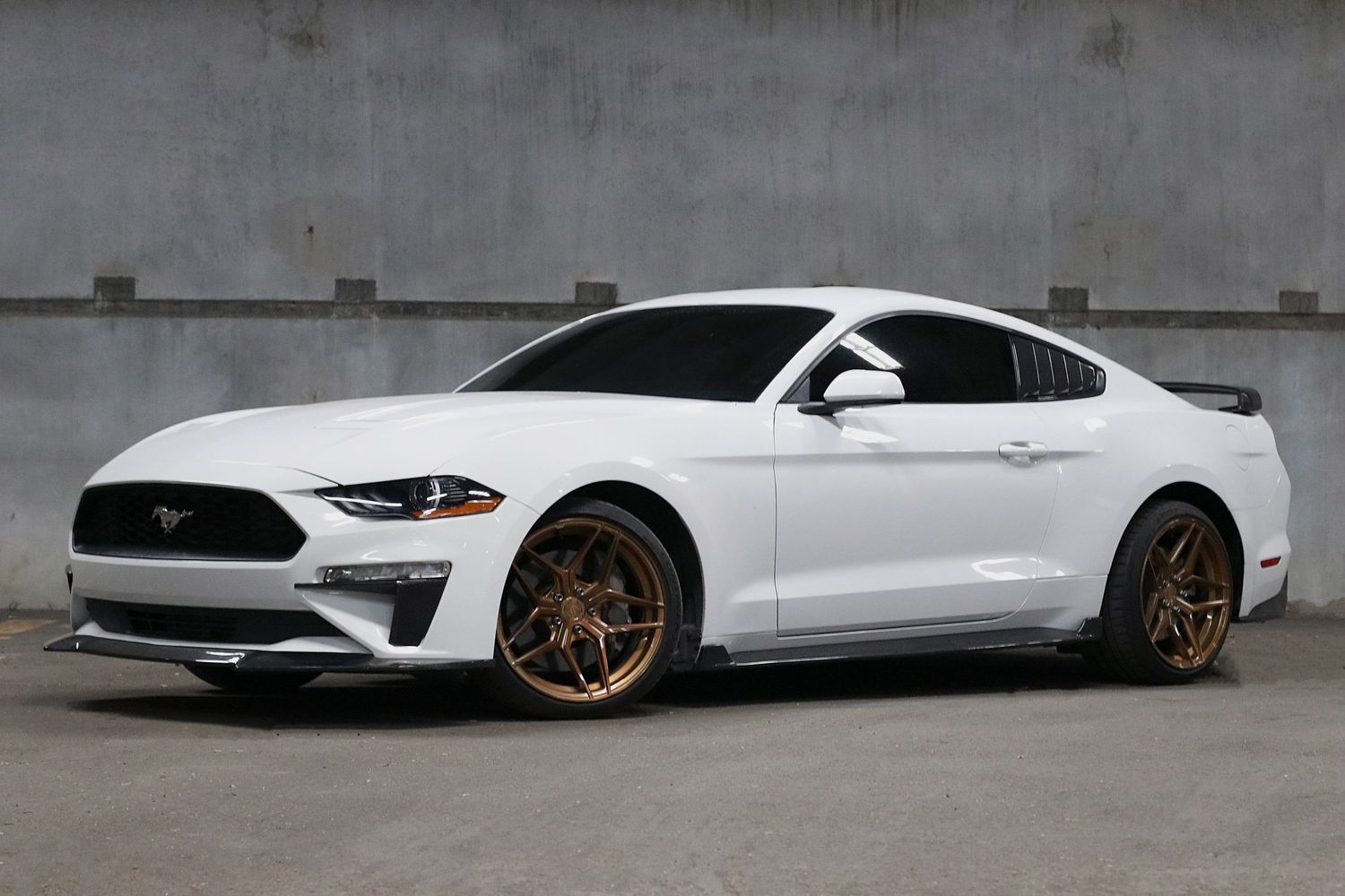 White Ford Mustang GT with gold aftermarket wheels parked in front of a concrete wall.