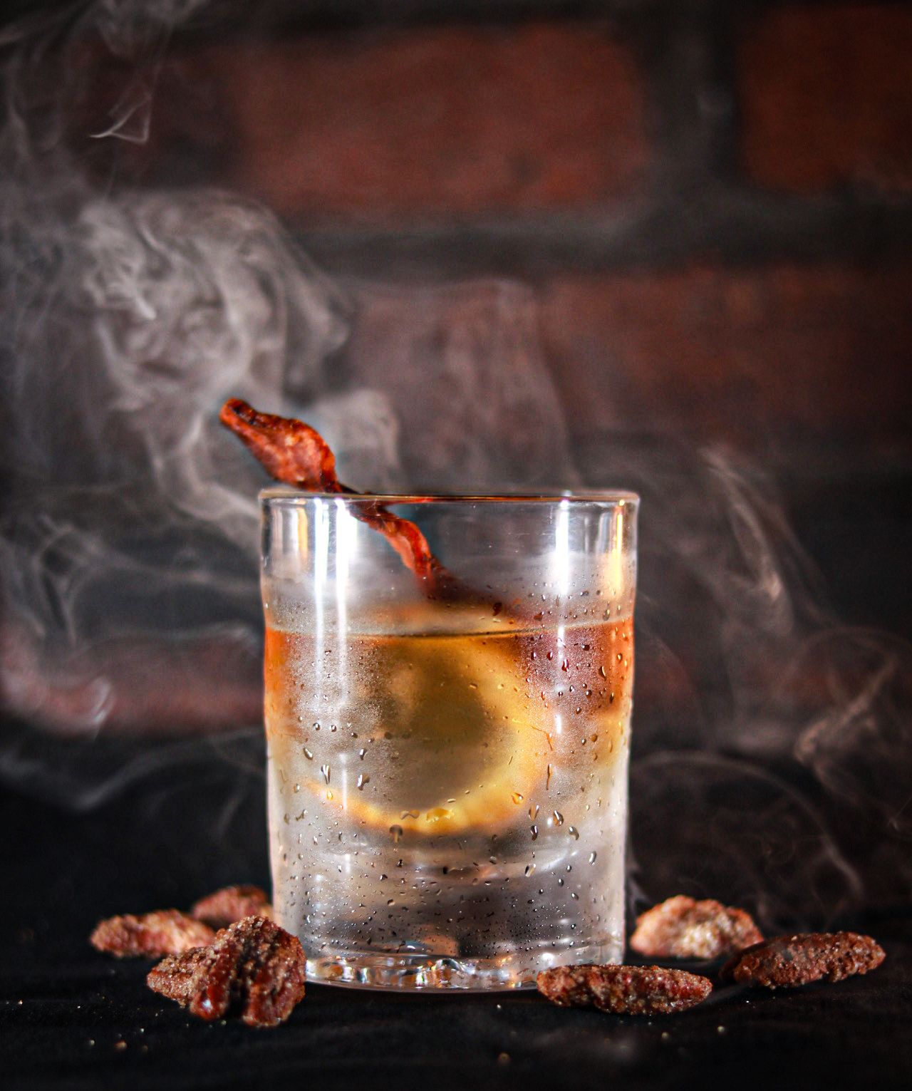 Smoky cocktail in rocks glass with candied bacon garnish, orange slice, and spiced nuts on dark surface.