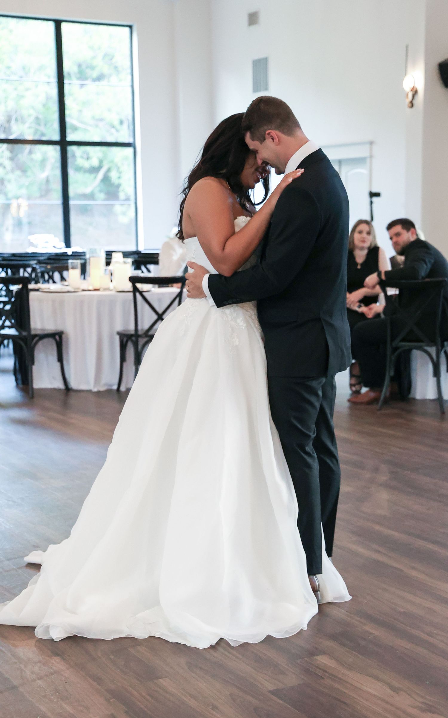 Bride and groom share their first dance in a bright wedding venue, embracing closely in formal attire.