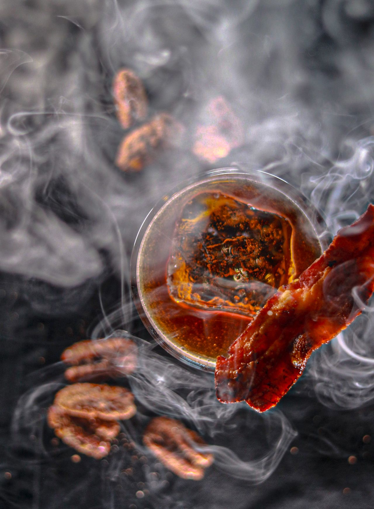 Smoky cocktail in glass with candied bacon garnish and spiced pecans surrounded by dramatic smoke swirls.
