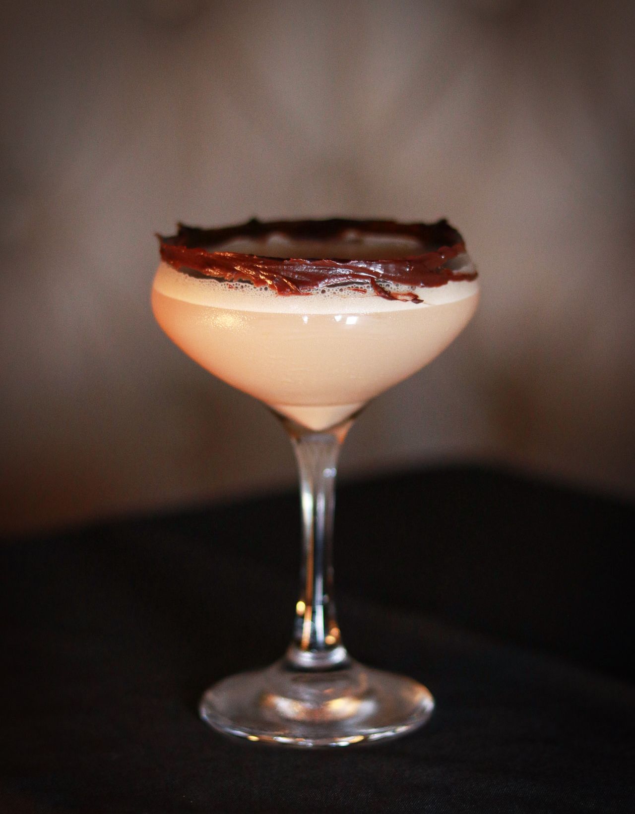 Elegant coupe cocktail glass with creamy drink, chocolate-rimmed garnish, on dark surface.