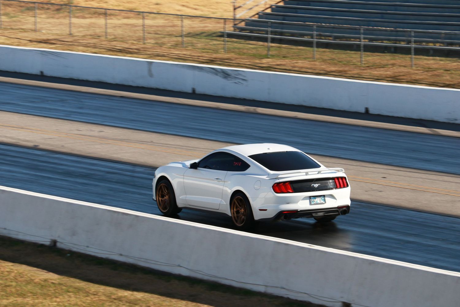 White Ford Mustang racing on a drag strip track with empty grandstands in the background on a sunny day.