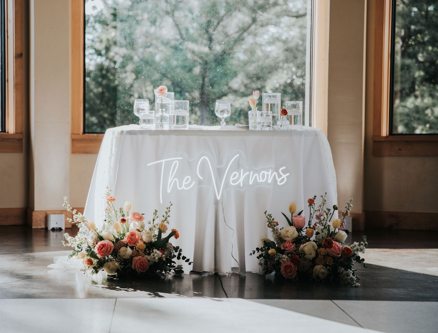 Elegant wedding sweetheart table with white linen, neon 'The Vernons' sign, and colorful floral arrangements by large windows.