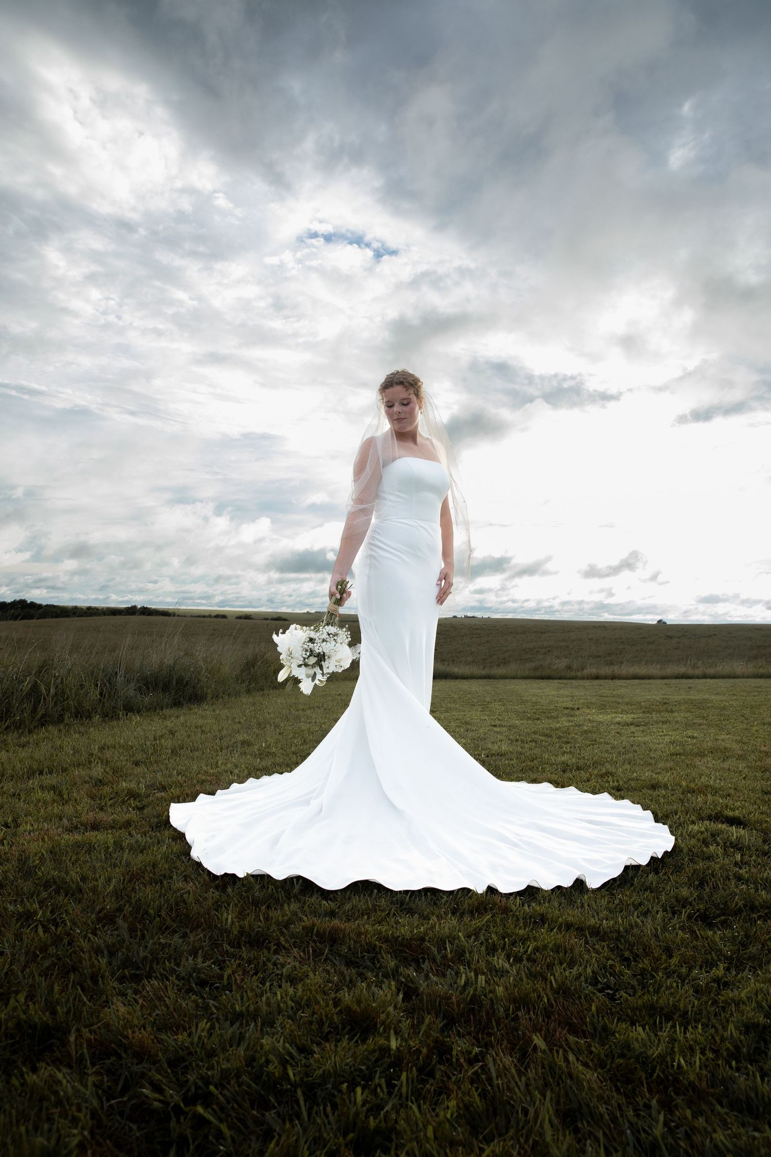 Bride in white mermaid gown with long train holds bouquet in green field under dramatic cloudy sky.