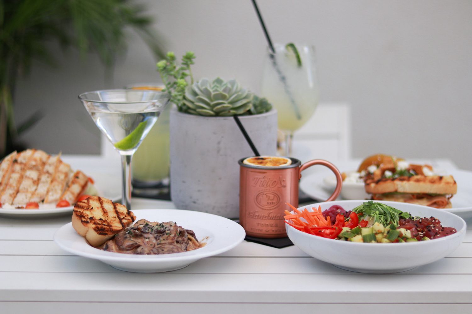 A spread of gourmet dishes and cocktails on a white table, featuring steak, salads, martini, and Moscow mule.