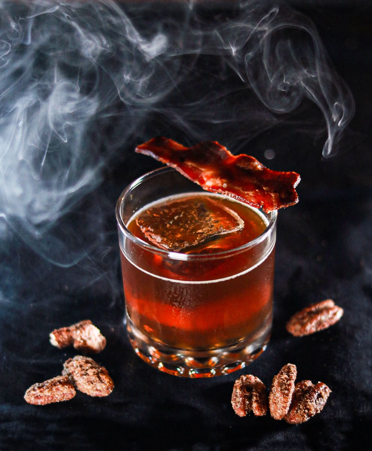 Smoky whiskey cocktail with bacon strip garnish, large ice cube, and candied pecans on dark background.