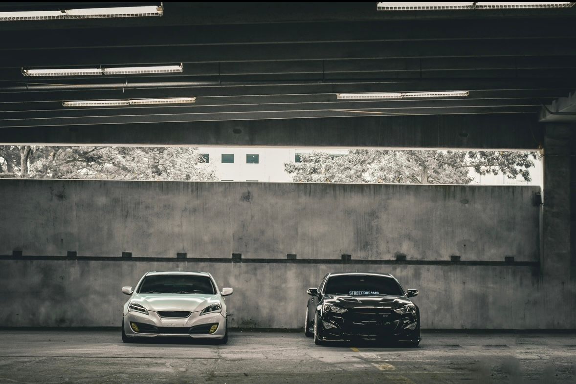 Two sleek modified cars, one white and one black, parked side by side in a concrete parking structure.