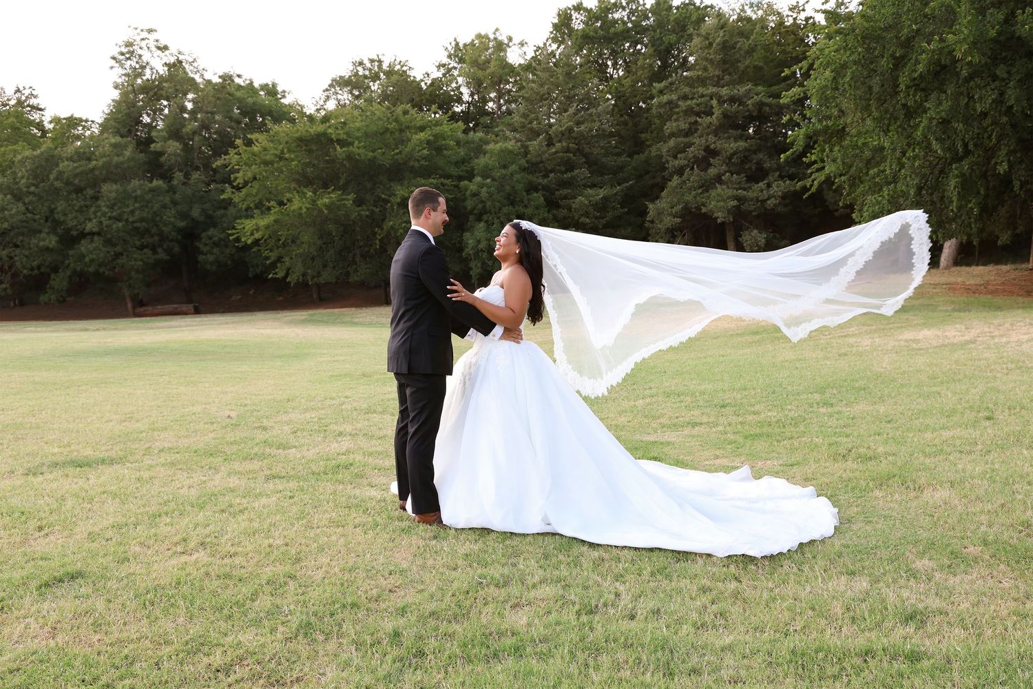 Bride and groom holding hands on green lawn, wedding veil flowing in wind, trees in background.