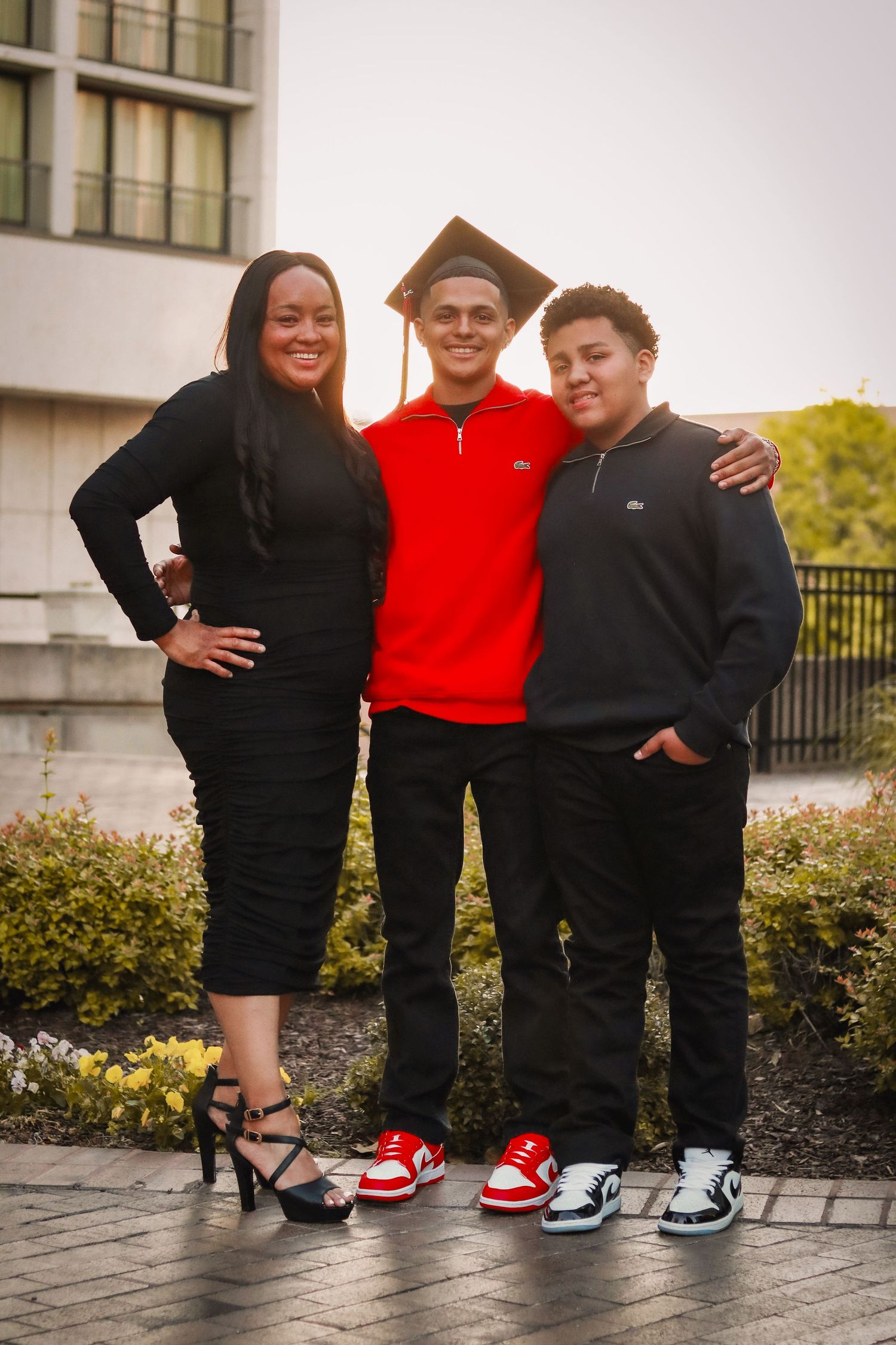 Three people stand together outdoors wearing casual attire including sneakers, with one wearing a graduation cap.