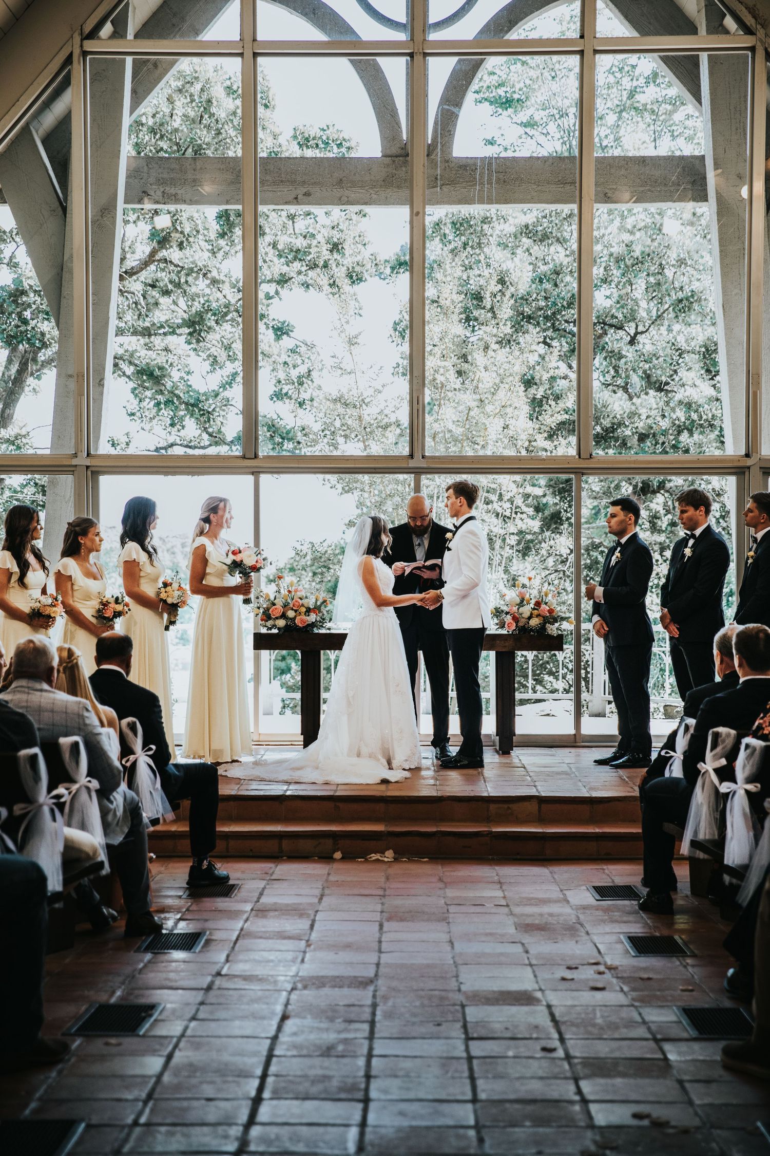 A bride and groom exchange vows at a stunning chapel altar with floor-to-ceiling windows and lush greenery backdrop.