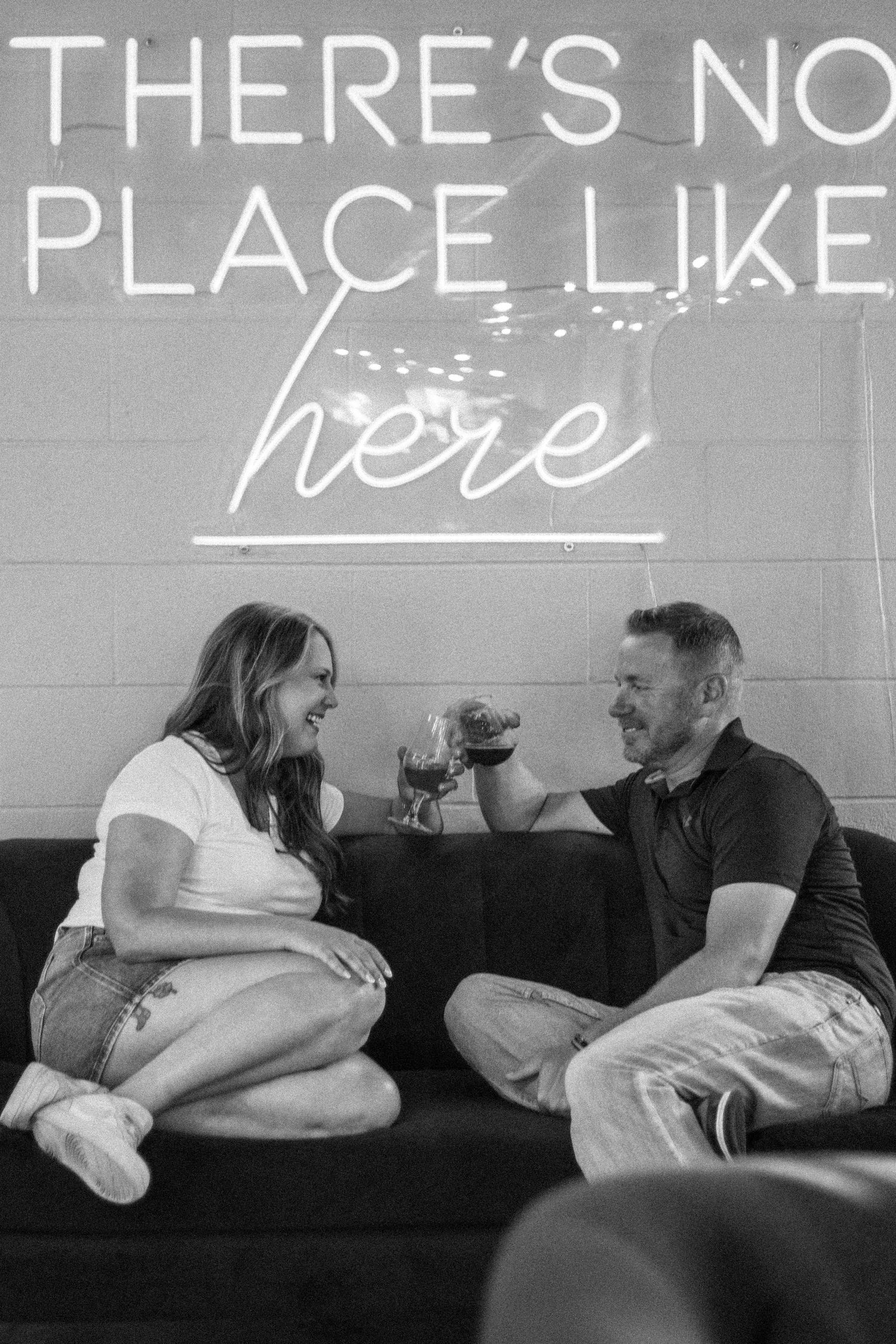Couple toasting with wine glasses on a couch beneath a neon sign reading There's No Place Like Here.