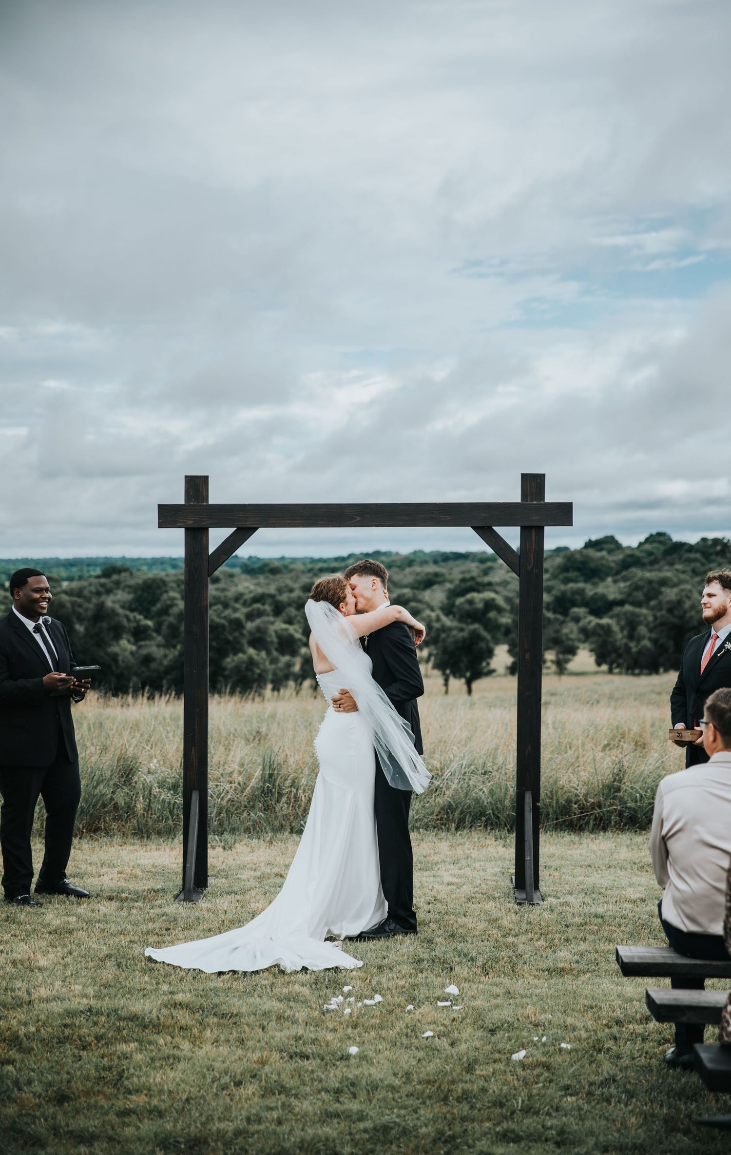 Bride and groom share first kiss under rustic wooden arch at outdoor wedding ceremony in open field.